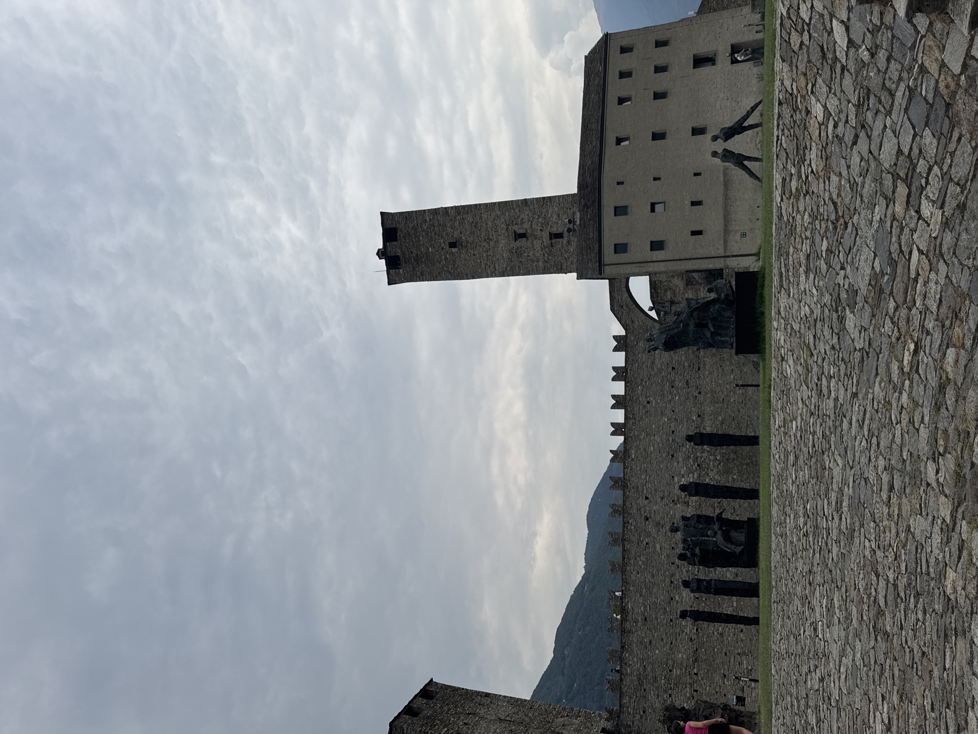 This is a full shot of the Castel Grande in Bellinzona, Switzerland, on an overcast day. The castle is made of stone and features several towers and battlements. In the foreground, there is a cobblestone courtyard. In the middle ground, there are several sculptures placed in front of the castle walls. In the background, there are mountains covered in trees. There are a few people visible in the image, mostly in the background. One person, wearing a pink top and black shorts, is standing on the left side of the frame.