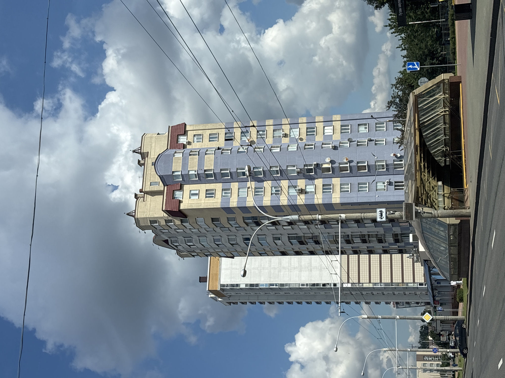 This is a medium-shot, eye-level, outdoor, daylight, vertical photograph of an urban street scene in Minsk, Belarus. In the foreground, a two-lane asphalt road runs horizontally across the frame, marked with white lane lines. To the right of the road is a modern glass-covered entrance to an underground passage or metro station. The entrance is illuminated by yellow safety markings on the floor and appears to be empty. A concrete pillar supporting power lines stands in front of the entrance, with a blue pedestrian sign showing a person walking upstairs mounted on it. Further to the right, a black trash receptacle is visible.
Dominating the midground are two tall, modern apartment buildings. The building on the left is slender and primarily white with horizontal brown bands and numerous windows. The building on the right is taller and wider, featuring a facade of alternating yellow and pale blue horizontal stripes. This building has a red-tiled, mansard-style roof with decorative dormer windows. Both buildings are adorned with multiple balconies, air conditioning units, and windows. Several streetlights and utility poles with hanging wires are positioned along the street, partially obscuring the view of the buildings.
In the background, lush green trees form a backdrop to the buildings. The sky above is a vibrant blue, filled with dramatic, billowing white clouds, some of which are large and dark, suggesting changing weather. A few smaller buildings and distant greenery are visible in the far background on the left. A dark-colored sedan is parked on the side of the road in the left midground. The overall impression is one of a well-maintained, modern urban environment under a dynamic sky.