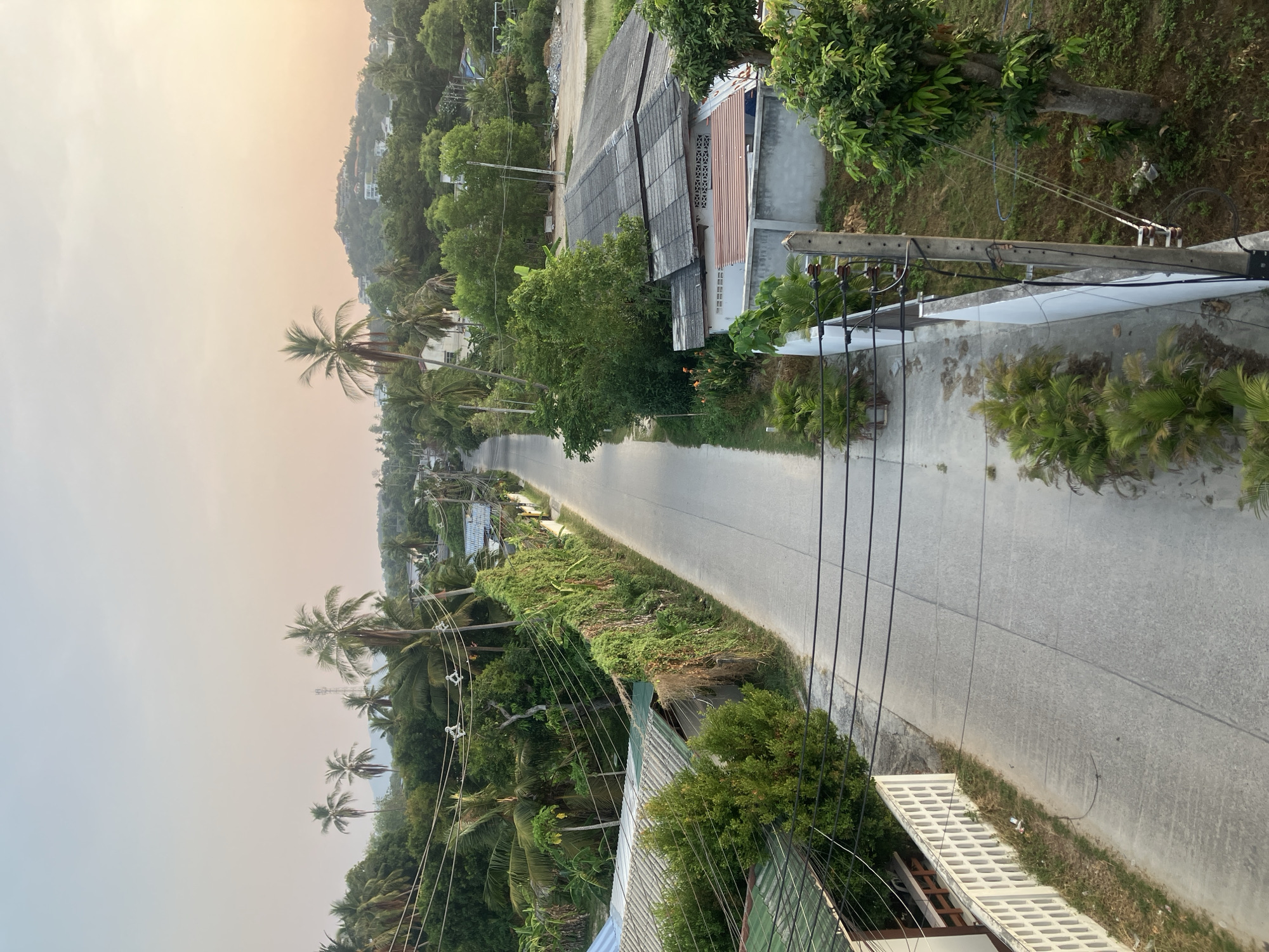 An aerial view depicts a straight road running through Baan Plai Laem, Thailand. The road is flanked by tropical vegetation, including palm trees. Buildings and houses with corrugated roofs line either side of the road. Utility poles and power lines stretch along the side of the road, running parallel to it. The sky is hazy with a gradient of light pink and orange.