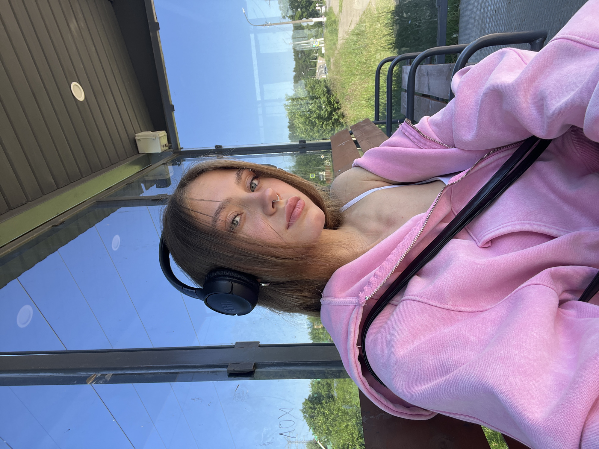A young woman with light brown hair, wearing black headphones, a white tank top, and an oversized pink zip-up hoodie, is sitting on a bench at a bus stop in Chaikovsky, Russia. She has a septum piercing and is looking towards the camera. The bus stop has glass walls and a grey, slatted roof. Outside, there is a grassy area with trees and a road, under a clear blue sky. The lighting suggests it is daytime.