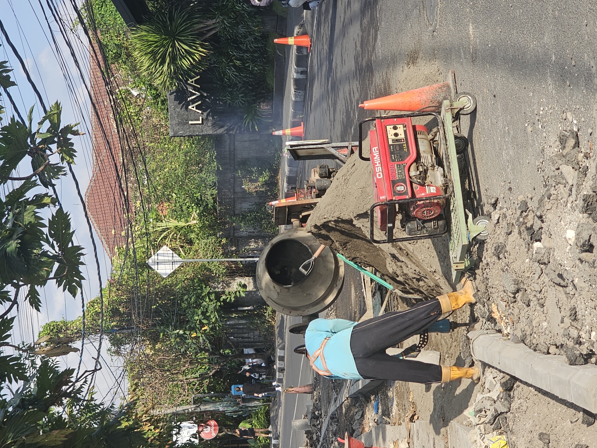 In the city of Denpasar, Indonesia, a construction worker, wearing a blue long-sleeved shirt, black pants, and yellow boots, operates a jackhammer on a street under repair. A large cement mixer stands nearby, filled with sand. A red portable generator with the brand "OSHIMA" is on a green trolley next to a pile of sand. Several orange traffic cones are placed along the road. In the background, a sign with the word "LAVA" is visible on a dark facade, surrounded by lush greenery and power lines overhead, suggesting a tropical setting. The scene appears to be a typical urban construction site.