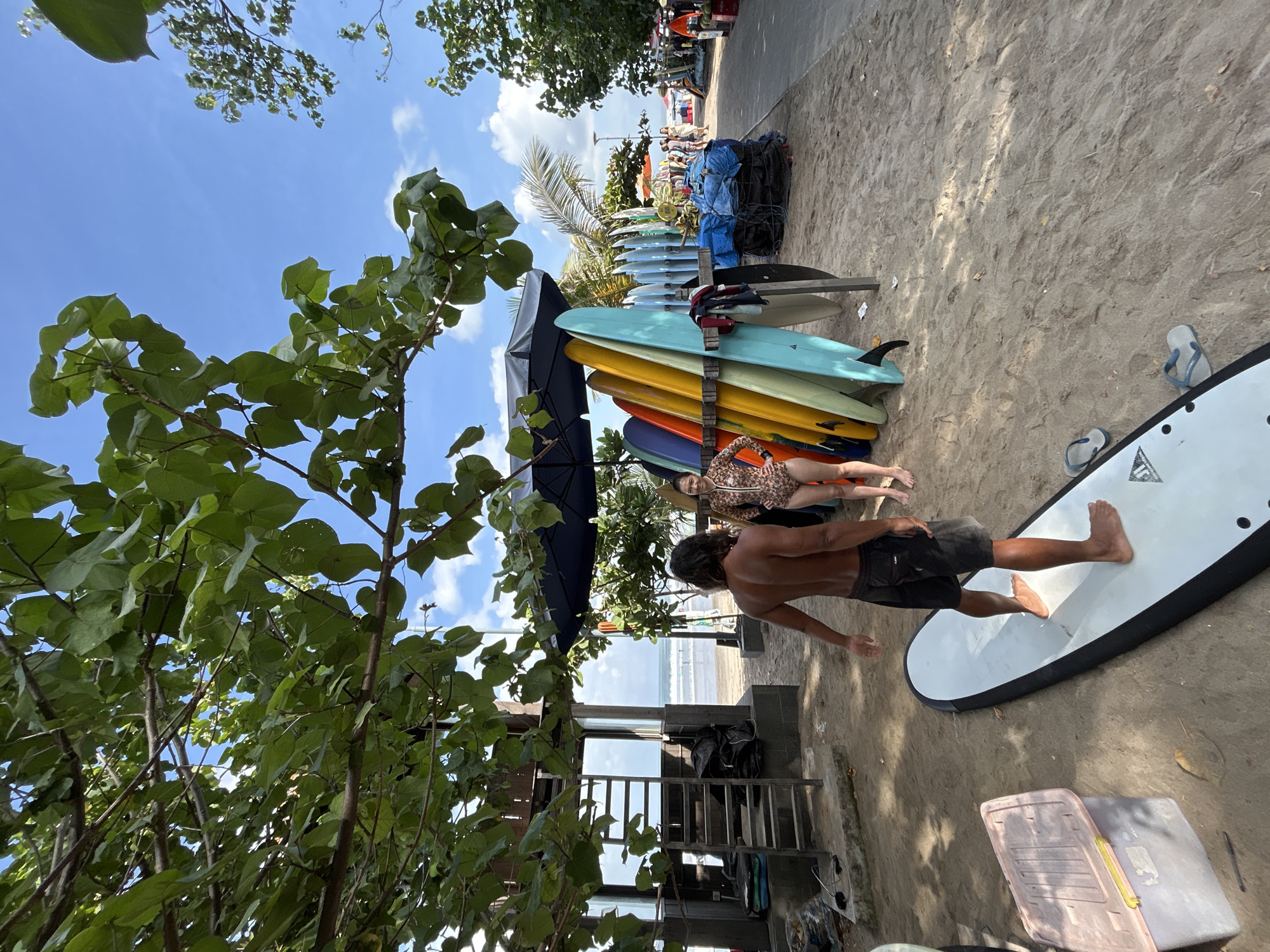 A man with long, dark hair and a muscular physique, wearing black swim trunks, stands on a white surfboard on a sandy beach. He is facing away from the camera, his arms extended slightly as if balancing. To his right, a woman in a patterned one-piece swimsuit smiles and stands near a rack of colorful surfboards. The surfboards are arranged vertically, with blues, yellows, oranges, and other vibrant colors visible. Behind them, a dark blue umbrella provides shade.
In the background, the ocean with gentle waves is visible, and a few people can be seen in the water. To the left, a wooden structure with a ladder leading up to it, possibly a lifeguard tower or rental shop, is present. Bags are stacked near the base of the structure. The ground is covered in sand, with some fallen leaves scattered around. A pink and gray plastic storage container is visible on the left side of the frame. The scene appears to be in Kuta, Indonesia, a popular tourist destination known for its beaches and surf culture, likely a surf rental or lesson area. The sky is blue with scattered white clouds, and lush green leaves from overhead trees partially obscure the view.