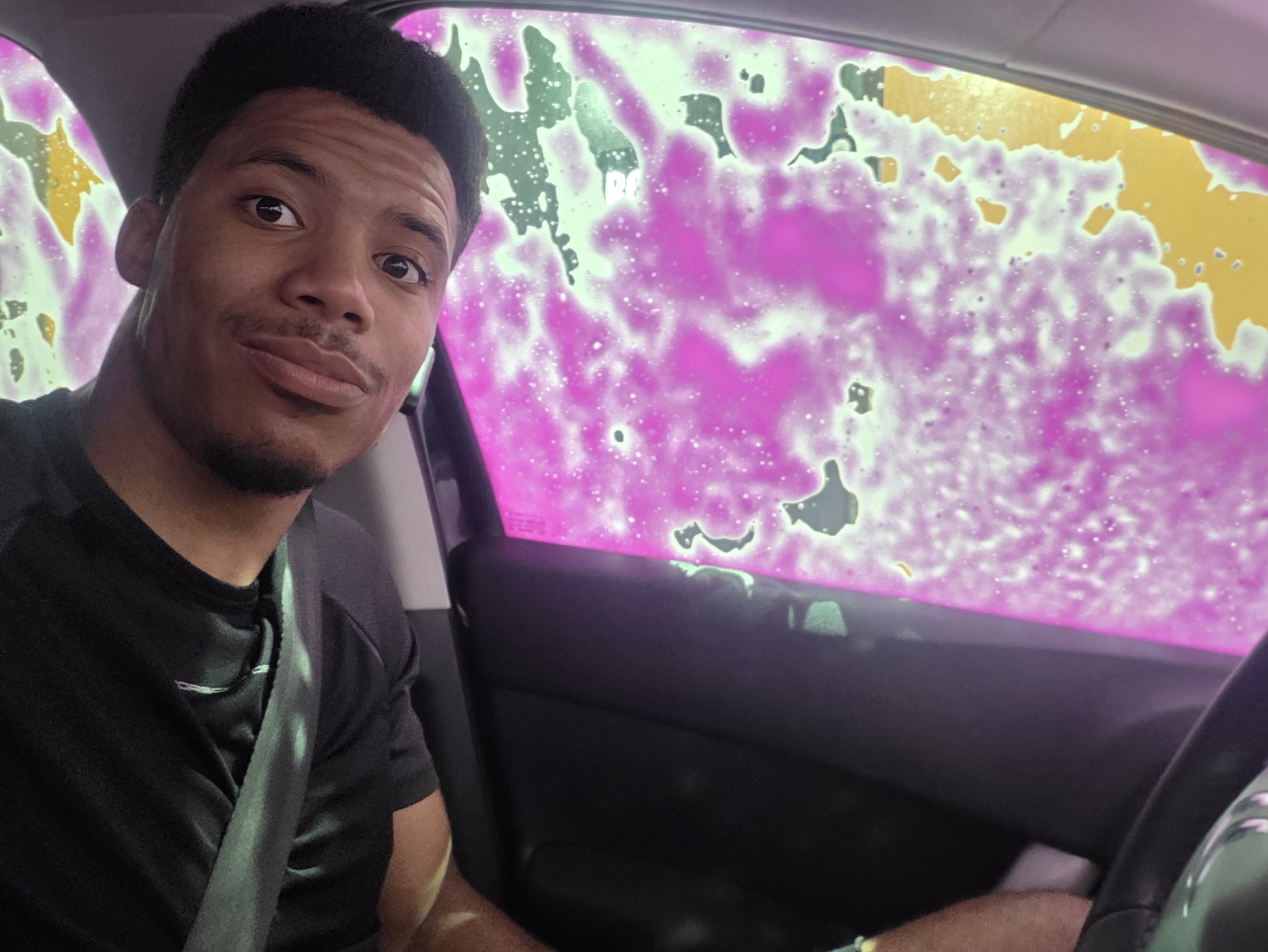 A young Black man is taking a selfie inside a car, possibly at a car wash, given the bright pink and yellow foam on the window. He is wearing a black t-shirt and a seatbelt. His left arm is visible, resting on the steering wheel. The background is obscured by the colorful foam. The lighting is bright and somewhat diffused. The overall scene is casual and personal. The car appears to be parked or moving slowly, likely in an urban environment given the context of a car wash. It is impossible to determine the precise location within Varna, Bulgaria, based solely on the visual information provided.