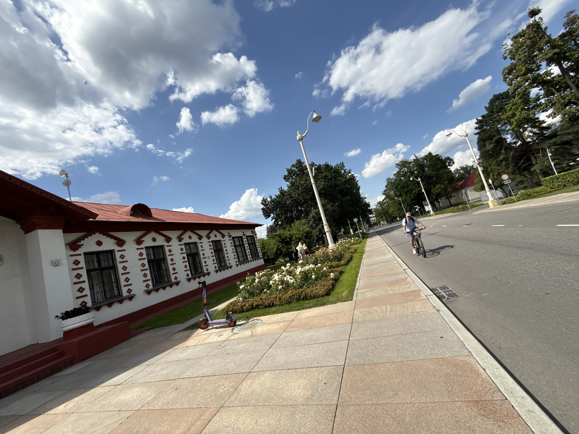 A fisheye lens captures a sunny day scene on a wide sidewalk next to a road in Moscow, Russia. On the left, a white building with a red roof and ornate trim features several dark-paned windows. In front of the building, a bed of white roses is in bloom, bordered by green grass and dark bushes. A purple electric scooter stands on the sidewalk near the building. Two people are seen walking near the flower bed. A man on a bicycle rides down the right lane of the asphalt road, casting a shadow. The sky is blue with scattered white clouds.