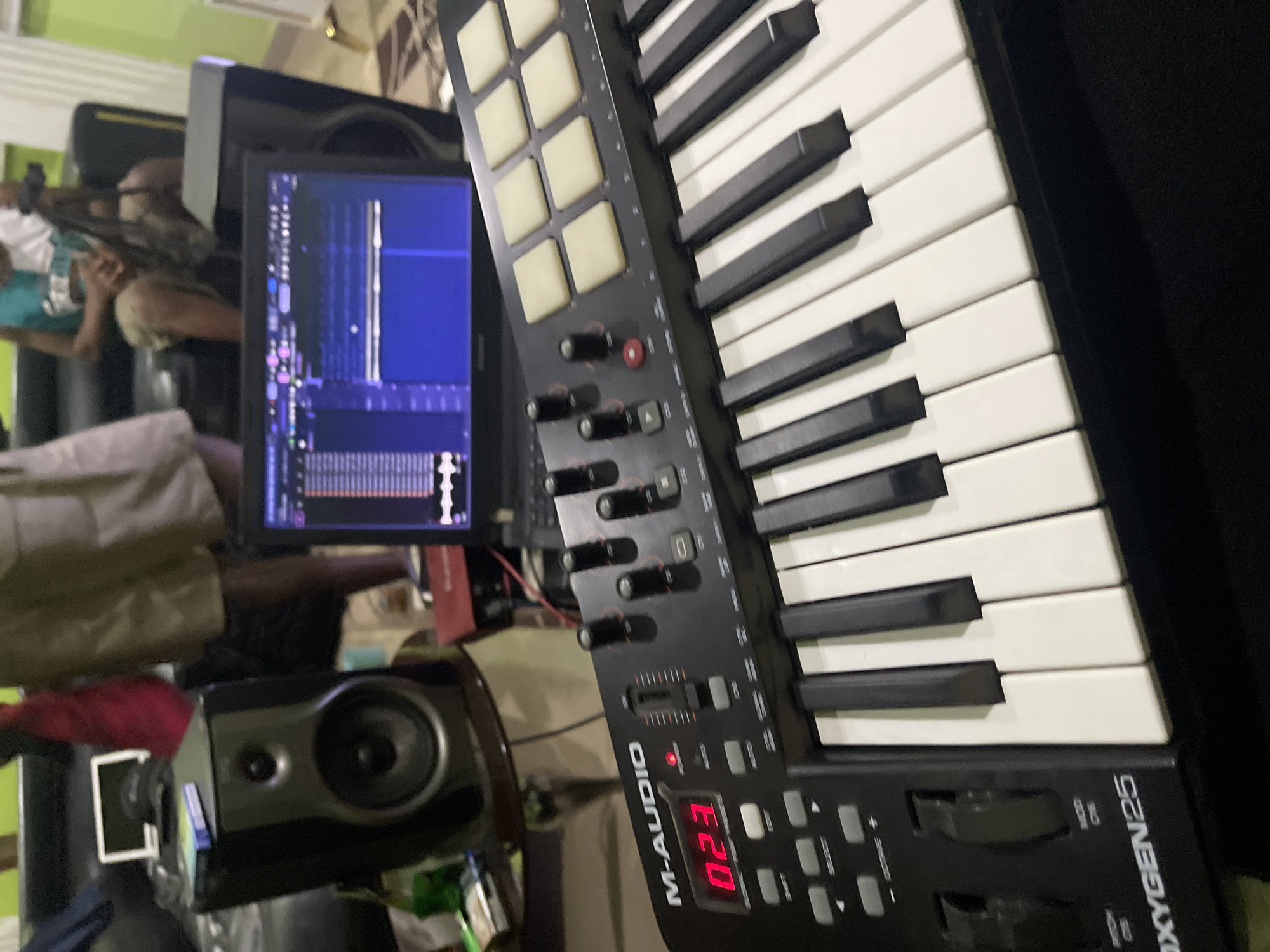 The image depicts a music production setup. In the foreground is an M-Audio Oxygen 25 MIDI keyboard, angled towards the right. Behind it is a laptop displaying music production software with waveforms visible on the screen. To the left of the laptop is a studio monitor speaker.
In the background, there are people seated on a dark-colored sofa. The environment appears to be an indoor space, possibly a home studio or recording space. It's not possible to independently verify that the photo was taken in Abuja, Nigeria, based on only the image content, although that was given as information. No city-specific identifiers are visible in the photo.