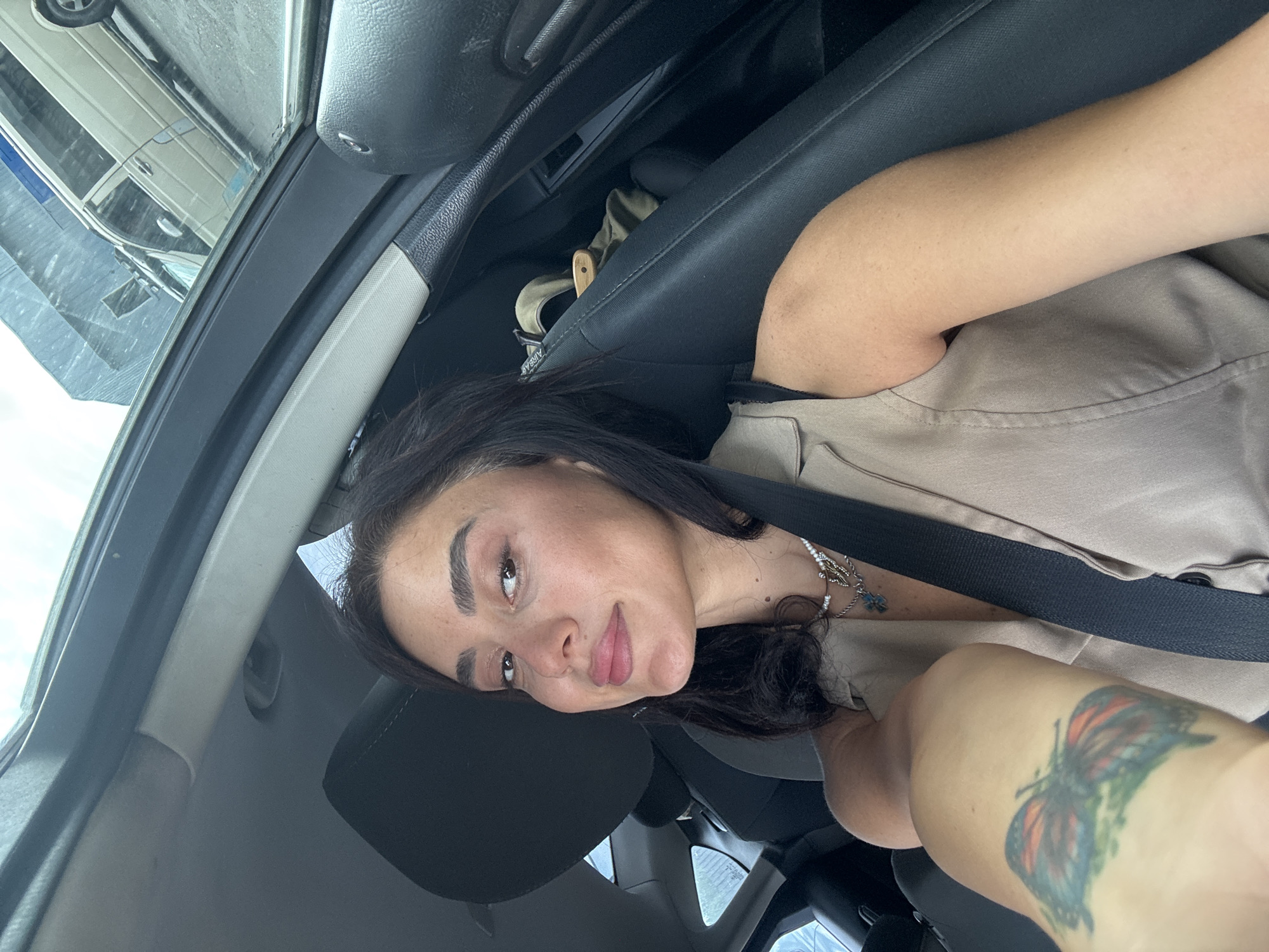 A medium shot captures a young woman with dark hair and brown eyes, wearing a beige sleeveless vest and a silver necklace, sitting in the driver's seat of a car. She is looking at the camera with a slight smile. A butterfly tattoo is visible on her left arm. The car's interior is dark, with black seats and a light-colored headliner. Outside the car, through the windshield, a light-colored van is visible, and reflections of buildings can be seen on the glass. The background is blurred. The image appears to be taken in an urban environment, potentially Tbilisi, Georgia, given the style of vehicles and architecture.