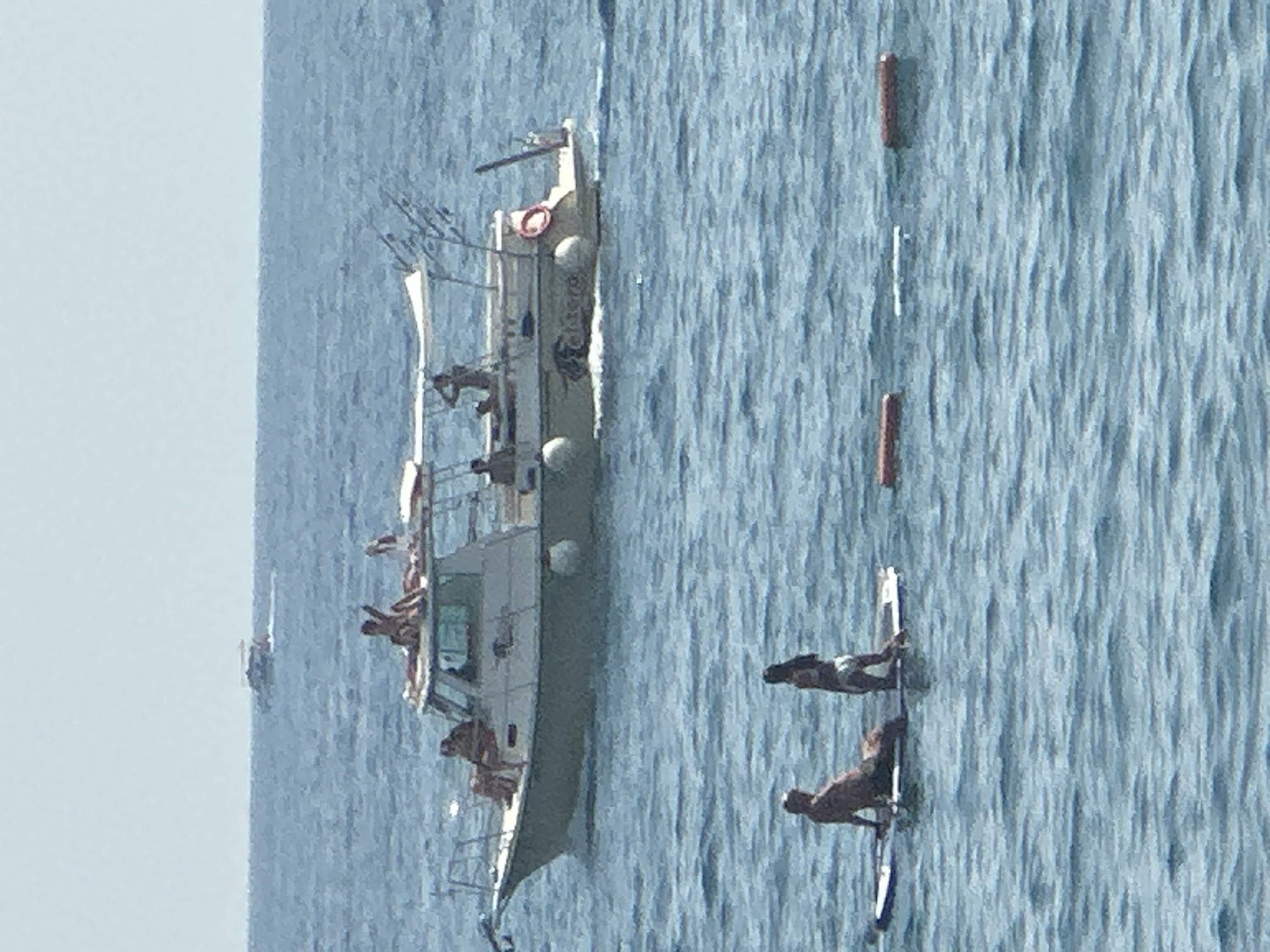 The image depicts a scene on the water near Boreti, Montenegro. In the foreground, two individuals are on a paddleboard. One person is lying down, and the other is kneeling. In the background, a white boat with a green hull is visible, carrying several people. The boat has a canopy providing shade. Further in the distance, another boat is moving across the water. The water is calm with small ripples. The sky is clear and light blue. Several buoys are scattered in the water.