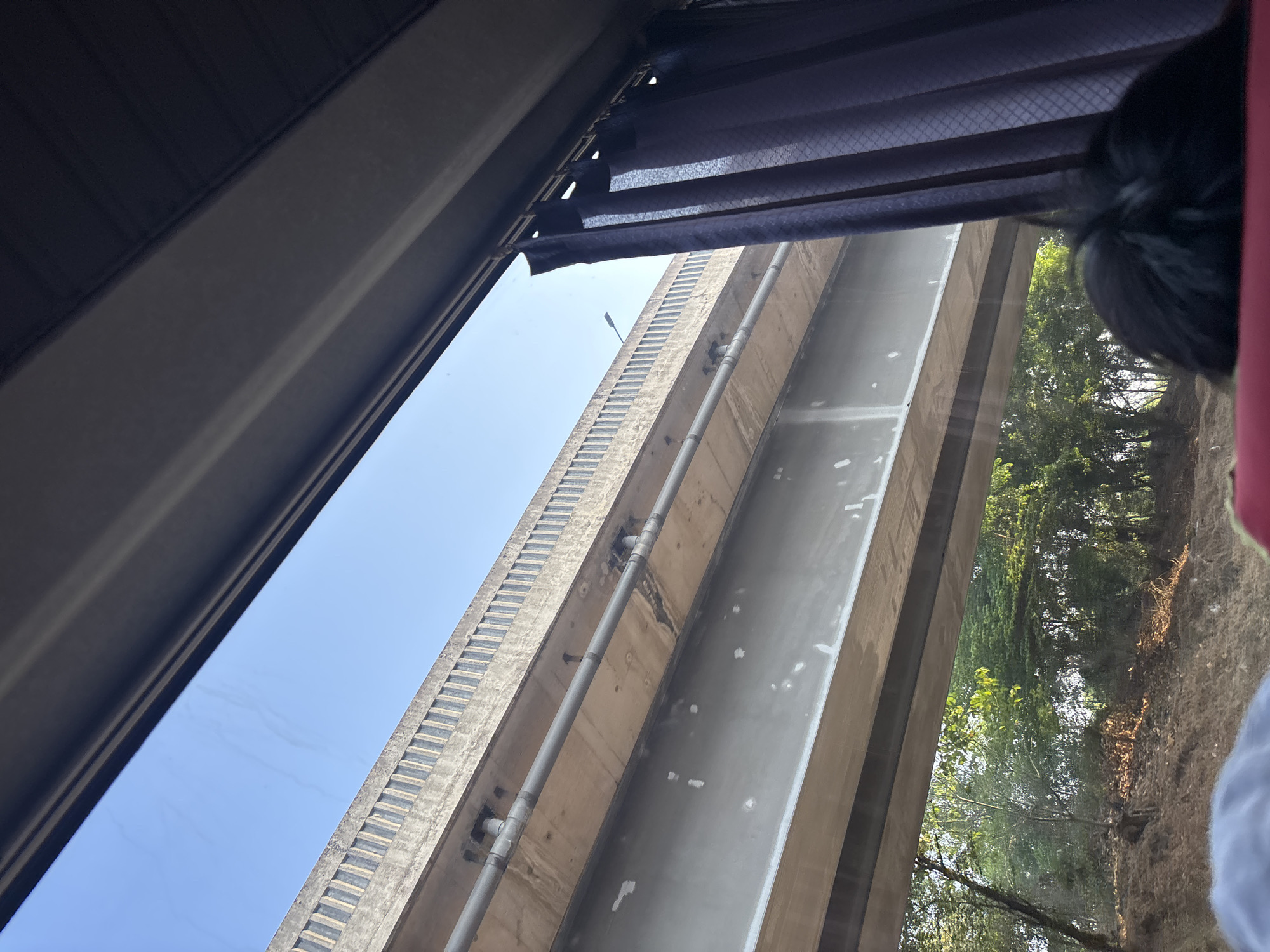 The image, taken from inside a vehicle in Karawang, Indonesia, shows a view through a window. A large concrete overpass dominates the upper portion of the frame, with a grey pipe running underneath it. The overpass has a decorative, textured concrete railing along its top edge. To the right, a dark purple curtain hangs down, partially obscuring the view. Below the overpass, a lush green forest can be seen with sunlight dappling through the trees. The foreground of the image shows the heads of two people, likely passengers in the vehicle, one with dark hair and the other with a blurry blue and grey object, possibly clothing, in the lower left corner. The scene suggests travel through an area with significant infrastructure and natural greenery.
