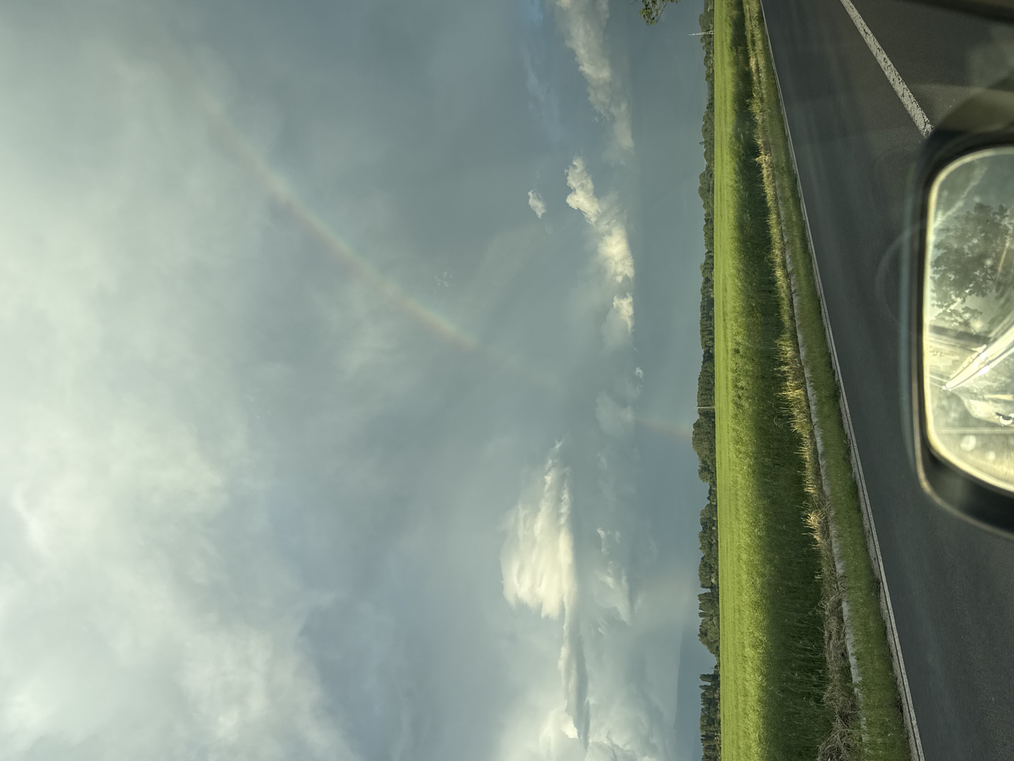 The image shows a view from inside a vehicle, likely a car, moving along a road. The road is paved with a white line marking the edge. To the right of the road is a green field or grassy area with a line of trees along the edge. In the sky, there are clouds, and a faint rainbow is visible. The rearview mirror of the car is visible in the right corner of the frame. The photograph was taken in the Gabarevo, Bulgaria, in the daytime.