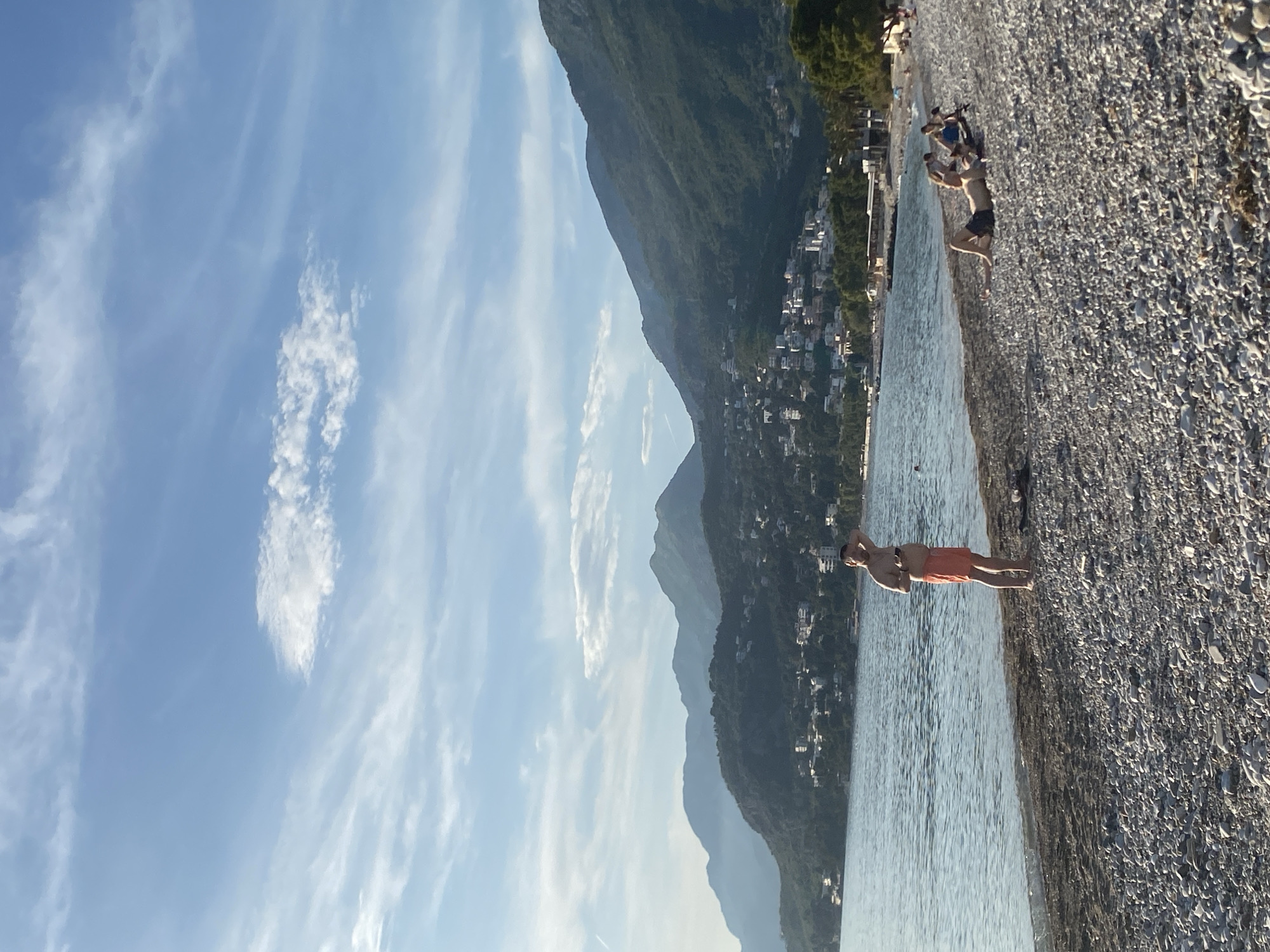 The image captures a sunlit coastal scene in Bar, Montenegro. The view features a beach composed of small pebbles, where a man wearing orange swimwear stands at the water's edge. Mountains rise steeply behind a row of buildings along the shoreline. The sky is blue with scattered white clouds.