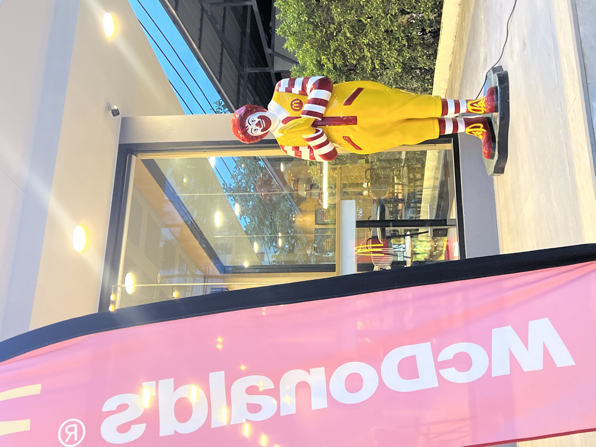 A sideways shot shows a McDonald's restaurant with a Ronald McDonald statue outside. The restaurant has a modern design with large glass windows reflecting the surrounding trees and sky. A bright pink McDonald's banner is in the foreground, partially obscuring the view. The statue of Ronald McDonald is posed with his hands clasped, and he is wearing his signature yellow jumpsuit, striped sleeves and socks, and red shoes. The environment appears to be a city setting.
