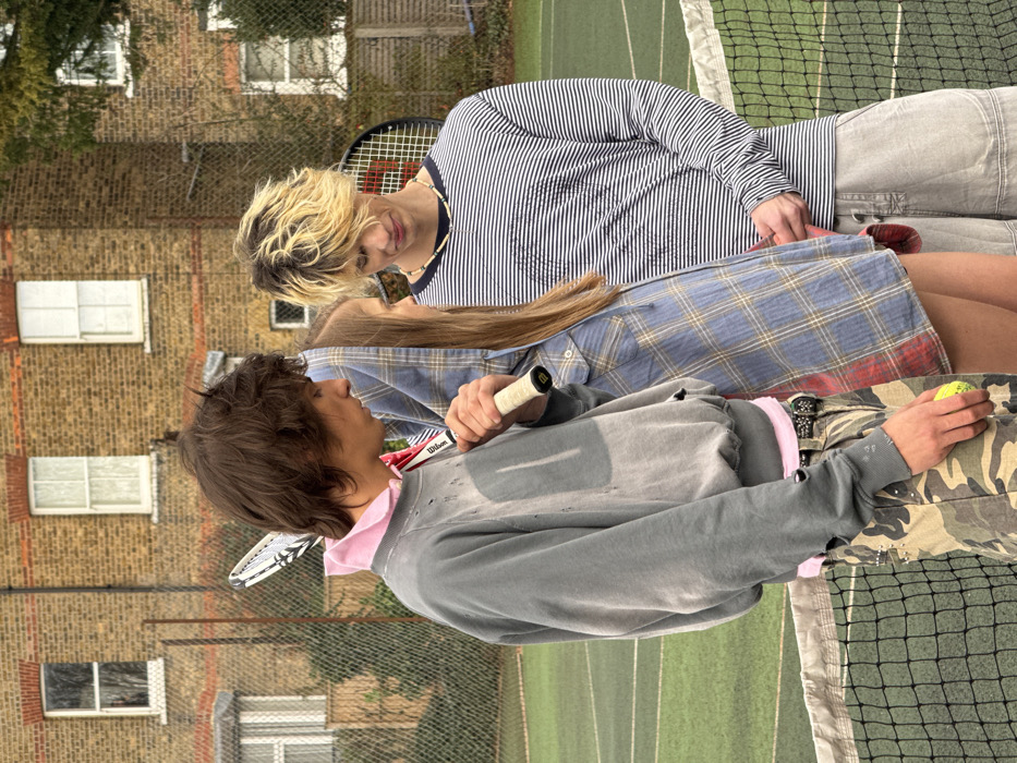 Two people stand on a tennis court, seemingly in conversation. One person wears a striped shirt and light-colored shorts, holding a tennis racket. The other is dressed in a gray sweatshirt over a pink collared shirt and camouflage pants, also holding a racket and a tennis ball. A tennis net is visible, along with a multi-story brick building in the background. The photo is slightly angled.