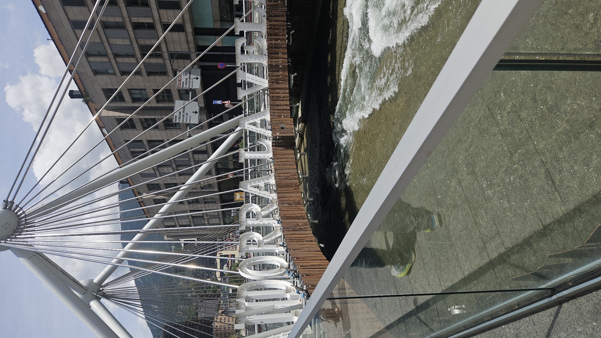 The image captures a detailed view of a modern pedestrian bridge in Andorra la Vella, Andorra, spanning a river with noticeable currents. The bridge features a distinctive white, cable-stayed structure, with numerous taut cables radiating from central hubs. Large, three-dimensional white letters spell out "ANDORRA LA VELLA" along the side of the bridge, positioned above a wooden paneling that forms part of the bridge's underside. The river beneath the bridge flows from left to right, exhibiting choppy waters and whitecaps. On the far bank, multi-story buildings with numerous windows line the river, backed by distant mountains under a partially cloudy blue sky. Several road signs are visible on the far side of the river, one indicating "ORDINO LA MASSANA" with directional arrows. In the immediate foreground, a glass and aluminum railing delineates the bridge's walkway. The wet pavement of the walkway reflects the lower legs and feet of a person wearing jeans and light-colored sneakers with neon yellow-green accents, suggesting the person is standing on the bridge and taking the photograph. The scene is illuminated by bright daylight, casting reflections on the bridge's cables and the wet ground.
