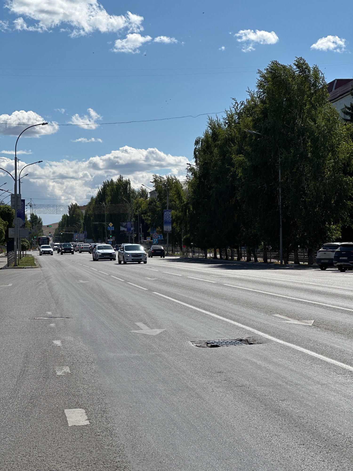 The image depicts a wide, multi-lane asphalt road in Kokshetau, Kazakhstan. The road is lined with tall, mature trees on the right side and on the left side in the distance. Several vehicles, including cars and a bus, are driving on the road. Traffic lights and streetlights are visible. The sky is blue with scattered white clouds. In the foreground, road markings, including white lines and arrows, are present. A manhole cover is visible in the road.
