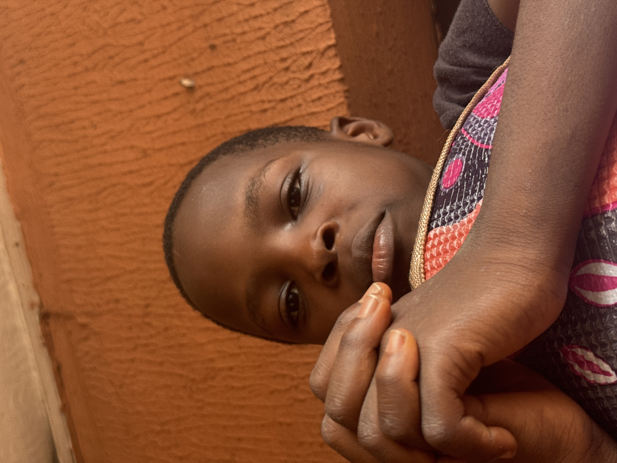 The image is a close-up, slightly angled shot of a young person's face, tilted and partially obscured by their arm and a patterned fabric. The subject's skin tone is dark, and their features are clearly visible, including their eyes, nose, and lips. Their hair is short and dark. The subject's hands are clasped together near their mouth, with one hand resting on their cheek.
The background is a textured, rust-colored wall, suggesting an outdoor or rustic indoor setting. A section of patterned fabric, featuring pink, white, and dark purple designs, is draped over the subject's arm and shoulder. The lighting appears natural, and there are no visible indications of the time of day or weather conditions. The overall mood is somewhat introspective, with the subject's gaze directed towards the viewer but with a contemplative expression.
The location context of Onitsha, Nigeria, suggests a setting within this West African city. The textured wall and the fabric could be common elements in the region. There is no visible text in the image.