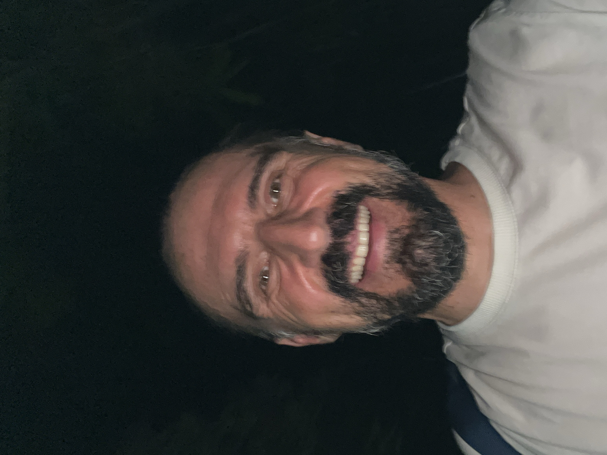 The image features a close-up, waist-up portrait of a smiling middle-aged man, with the original photo appearing rotated 90 degrees clockwise. The man has fair skin, a full beard mixed with grey and dark hairs, and short dark hair on his head with visible grey strands. He is looking towards the camera with a broad, joyful smile, showing his teeth, and his eyes crinkle at the corners, conveying amusement or happiness. He is wearing a white, long-sleeved top, possibly a sweatshirt, with a ribbed collar. A dark blue or black strap or piece of clothing is visible below his right arm.
The background is very dark and indistinct, suggesting the photo was taken at night or in a very dimly lit environment. The man's face is brightly illuminated, likely by a direct light source or flash, creating highlights on his skin and contrasting sharply with the dark surroundings. No specific objects or landscape features are discernible in the background. The man appears to be posing for the photograph in a casual, happy moment. The location context provided is บ้านบางรักษ์, Thailand. There is no visible text in the image.