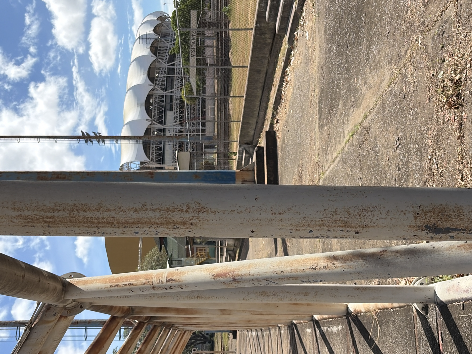 The image shows a view through rusted metal railings of a concrete structure. The railings are painted white and are showing signs of rust and peeling paint. Beyond the railings, there's a concrete area and a tall stadium-like structure with a white arched roof. The structure is labeled "VISITANTE". The sky is visible in the background with fluffy white clouds. It is most likely taken at the Cachamay Sports Center in Ciudad Guayana, Venezuela. The image appears to be taken at ground level, looking through a pedestrian walkway. There are no people visible in the image.