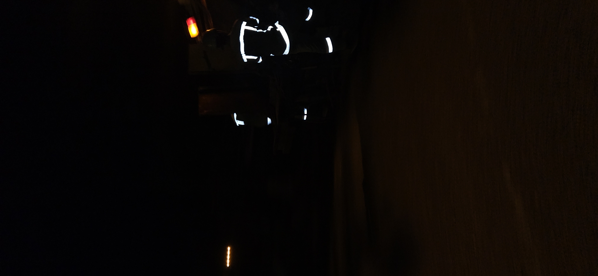 The image is extremely dark, predominantly composed of deep shadows, suggesting a night scene or a very dimly lit indoor or enclosed space. The primary subject is a person, or possibly multiple individuals, identifiable solely by bright, white reflective strips on their clothing, indicative of safety or work attire. These strips appear on the torso, shoulders, and potentially limbs, forming distinct, often curved, segments of light against the darkness.
To the upper left of these reflective elements, a clear red light is visible, which could be part of machinery, a vehicle's tail light, or an environmental indicator. Another small, vertical, orange or yellow light streak is present near the bottom center of the frame, possibly a reflection or a small light source on the ground. The right side of the image features a dark, textured surface that resembles a wall.
Due to the limited visibility, the specific activity or event taking place is unclear, but the presence of reflective gear suggests work, safety, or passage through a low-light environment. There are no discernible weather conditions or explicit location cues within the image itself. No text is visible.