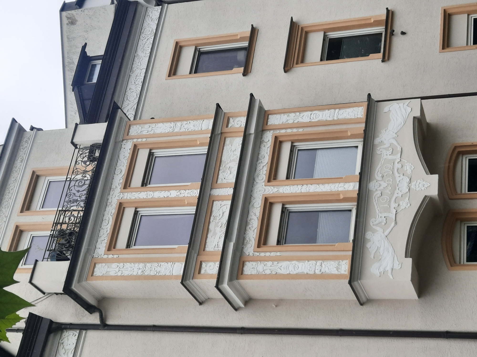The image shows a section of a light-colored building with ornate architectural details. The building has multiple windows, some of which are bay windows with decorative trim in peach and white. There is a small balcony with a wrought-iron railing on the upper level. A downspout runs vertically along the left side of the building. The sky is overcast. The architectural style suggests it could be a residential building.
The building is located at Max-Bamberger-Straße 2 in Rosenheim, Germany. This is a historic residential building with Art Nouveau elements, dating back to the early 20th century. The stucco work on the facade is characteristic of the period.
