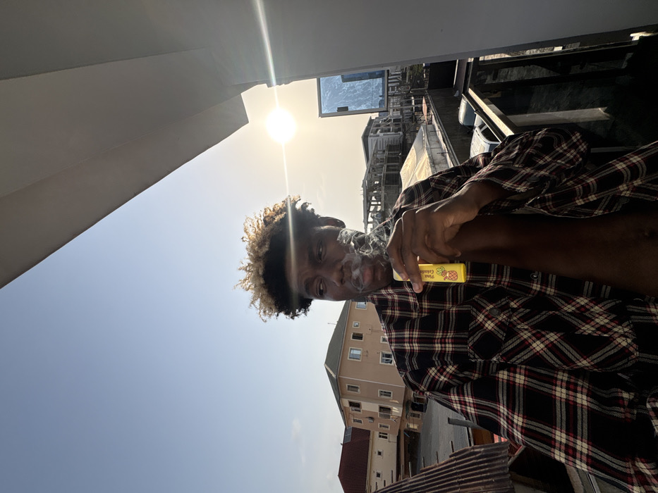 The image captures a male individual on a balcony in Igboefon, Nigeria. The subject, a dark-skinned male with short, curly hair featuring blonde highlights, is depicted from the chest up, wearing an unbuttoned black, red, and white plaid shirt. He holds a yellow, rectangular vaping device, branded "Pina Colada" with a pineapple graphic, in his right hand. A plume of white vapor is visible emanating from his mouth. His head is turned slightly, with his gaze directed toward the left of the frame. The background features a clear, light blue sky with the sun visible as a bright circular light source on the right, creating a lens flare. An urban landscape is visible below the horizon, comprising multiple multi-story buildings; one prominent structure to the left has a light brown facade and a dark red roof. Other buildings, some appearing to be under construction, are also present. Corrugated metal roofing is visible on the left side of the frame, and a dark metal balcony railing is in the immediate foreground.