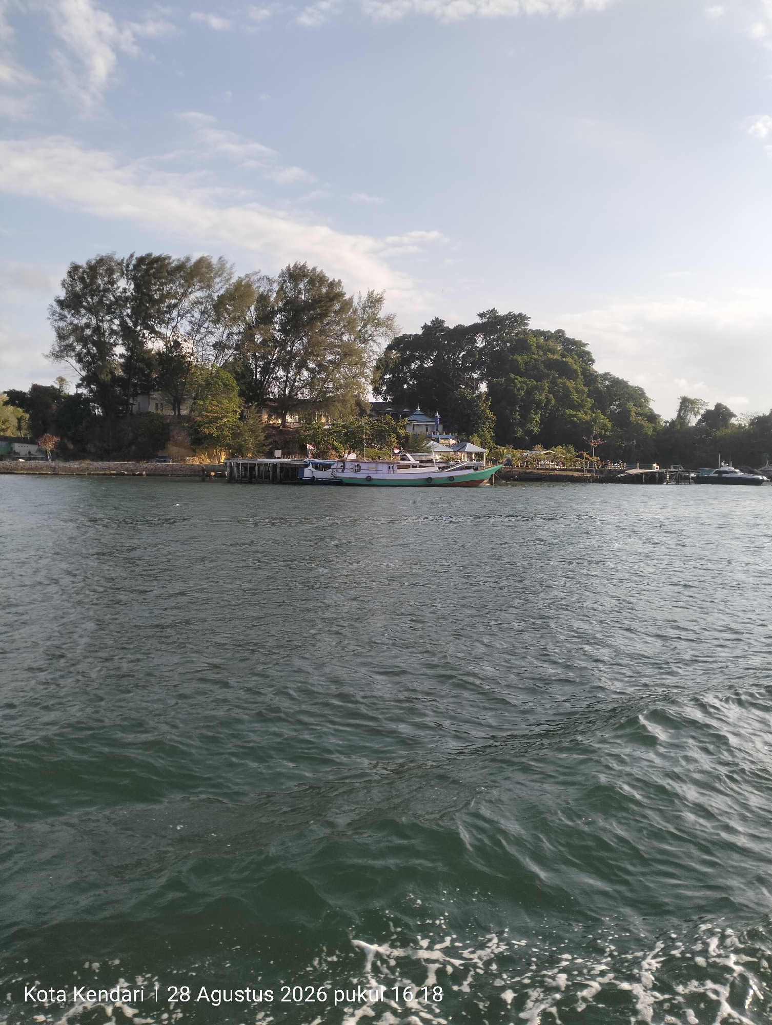A wide, calm body of dark green water with whitecaps in the foreground suggests movement, possibly from a boat. In the background, a tree-lined shore with a few buildings and a dock is visible. A green and white boat is docked at the pier. The sky above is light blue with scattered white clouds. The timestamp "Kota Kendari | 28 Agustus 2026 pukul 16:18" is superimposed on the lower left of the image.