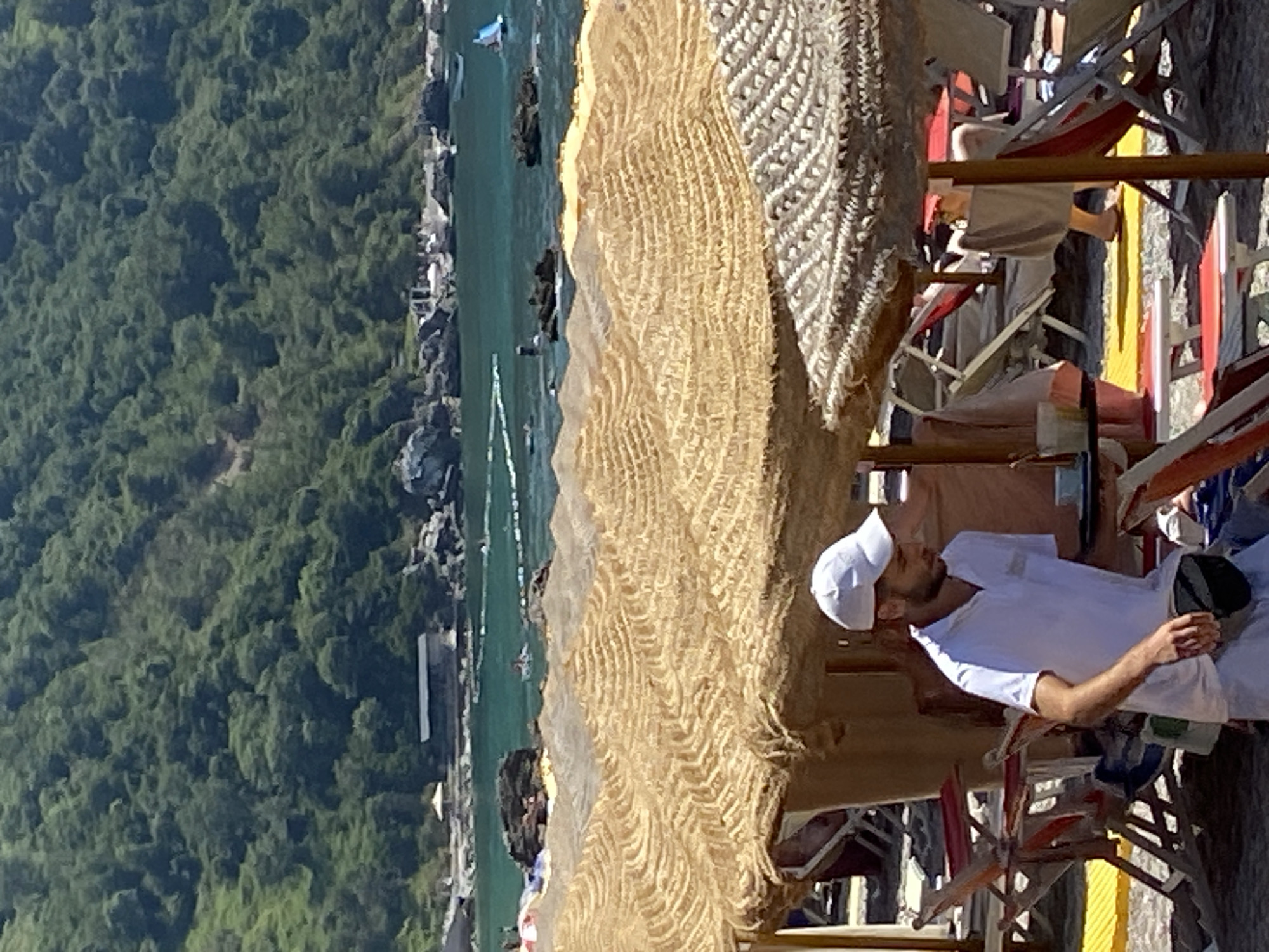 The image depicts a beach scene in Prijevor, Montenegro. In the foreground, a man wearing a white polo shirt and a white cap is holding a tray with a cup and a small bottle. He has a dark fanny pack around his waist. Behind him, several beach chairs and straw umbrellas are visible. The background shows the turquoise sea, a rocky coastline, and a lush green hillside with some buildings. Several people are scattered across the beach and in the water.