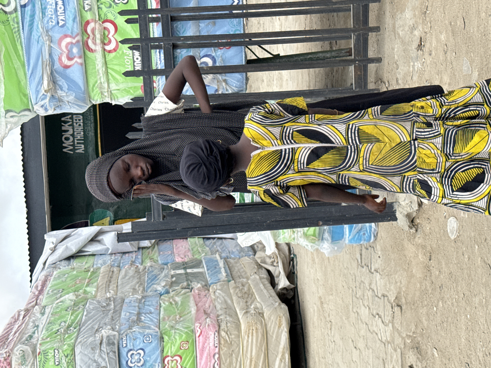 Two individuals stand near a street-side vendor selling mattresses in Lekki, Nigeria. One person wears a black and white dotted headscarf and a dark patterned top, while the other is in a yellow and black patterned dress. Stacks of mattresses with varying colored floral patterns are visible, and signage indicates the mattresses are by Mouka. The setting appears to be a busy street with a concrete surface.