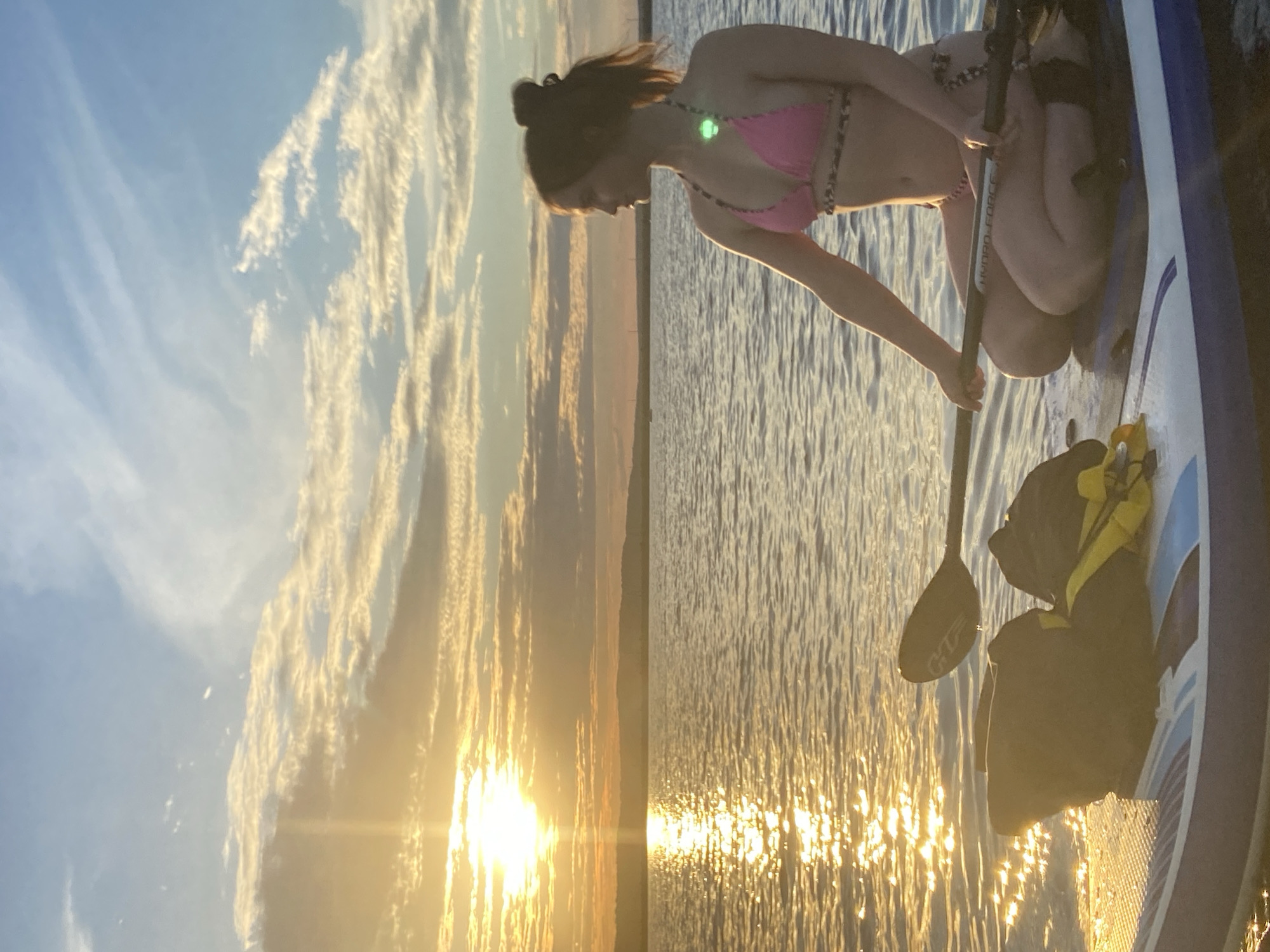 A woman in a pink bikini with a leopard print trim is on a paddleboard in the water. She is holding a paddle and appears to be preparing to move or has just finished paddling. The sun is setting, casting a golden glow on the rippling water, creating a beautiful reflection. The sky is blue with streaks of white clouds, suggesting it is a clear evening. The woman has her hair tied up in a bun. A yellow and black life vest is secured to the paddleboard behind her. The paddle has text on it, appearing to read "HYDRO-FORCE". The image captures a serene moment of outdoor recreation on the water during sunset.
