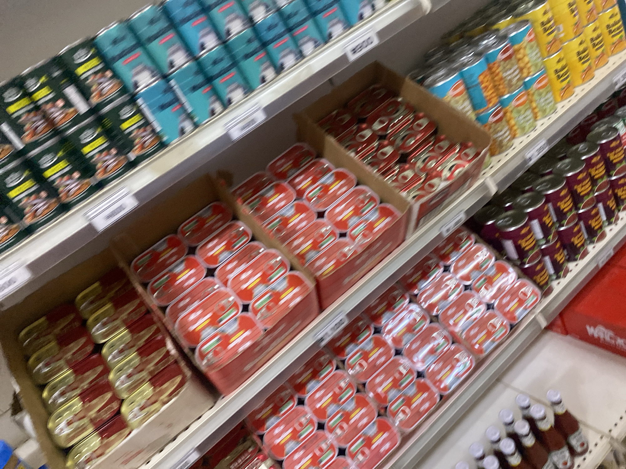 The image shows a shelf in a store, likely a supermarket or a convenience store, located in Ikorodu, Nigeria. The shelves are stocked with various packaged food items and drinks. On the top shelf, there are rows of canned goods, some with red and gold packaging, possibly sardines or other preserved food. Below this, another shelf displays more canned items in red packaging. Further down, there are bottles of what appear to be sauces or condiments, with brown liquid and white caps. The bottom shelves show more canned goods, with some featuring green and yellow packaging, and others with red and metallic labels. The overall environment appears to be indoors, with artificial lighting. The perspective is from an elevated angle looking down at the shelves, suggesting the photographer is standing or on a slightly higher level. The arrangement of products indicates a retail setting where items are displayed for sale.