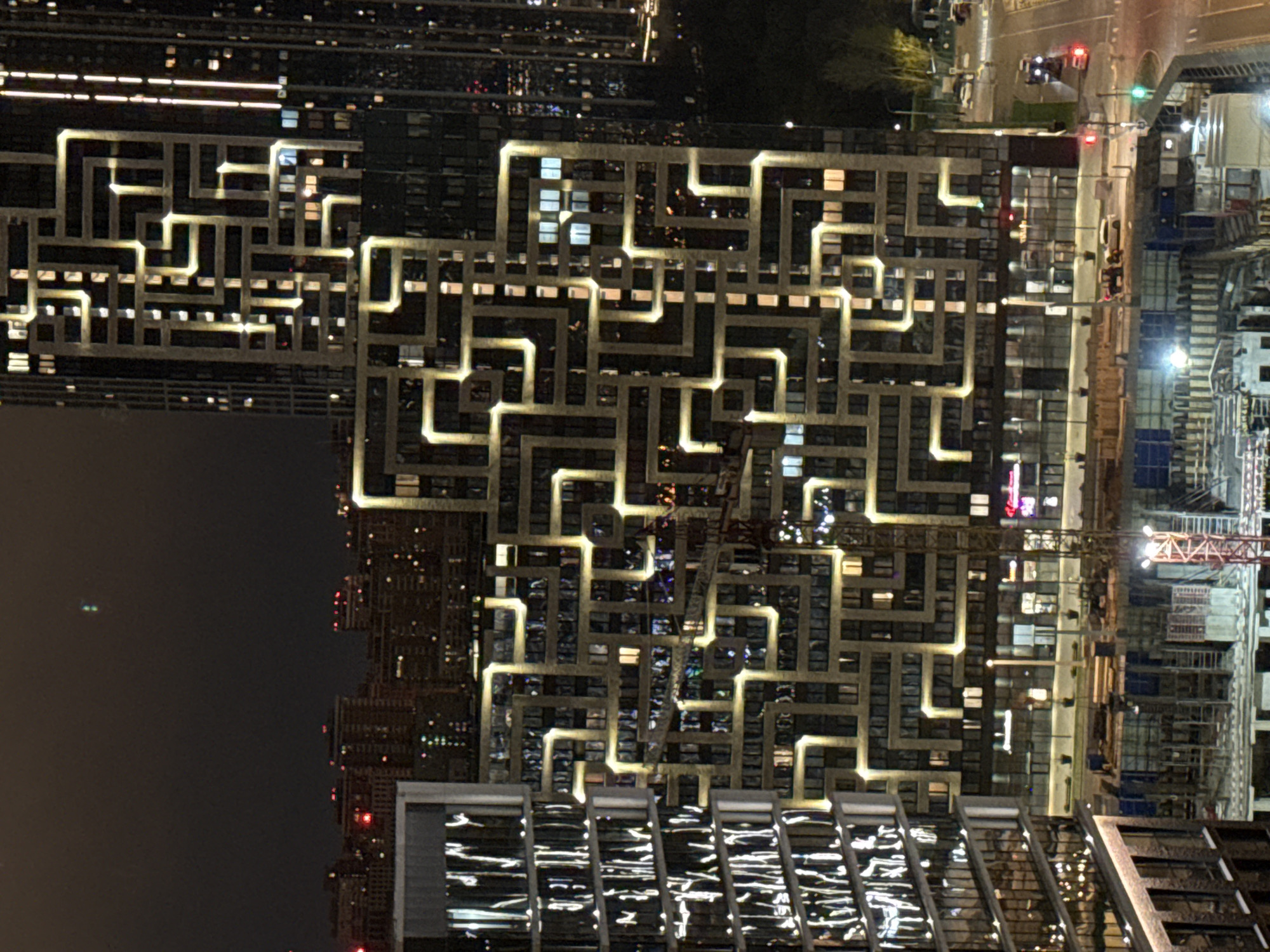 At night, a high-angle shot presents a view of buildings in Khimki, Russia. The most prominent building features an illuminated, maze-like design on its facade. To the right, a construction site includes a crane and building materials. Streetlights and vehicle lights mark a road in the background. Other buildings are visible in the distance.