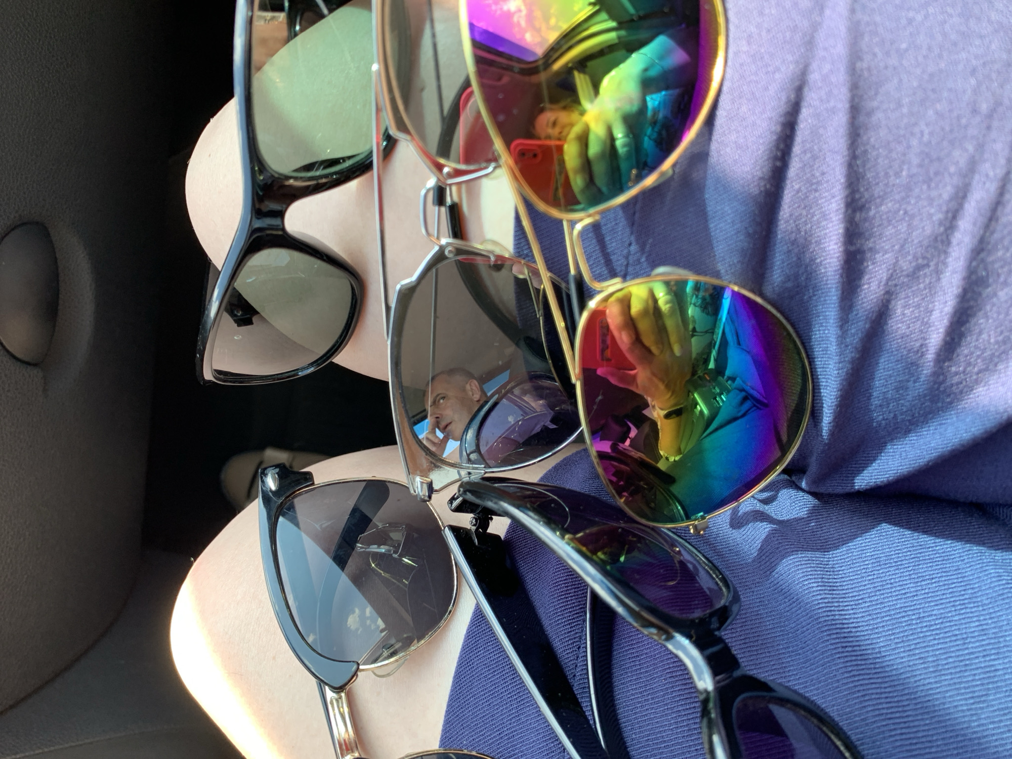 A close-up, overhead shot shows several pairs of sunglasses on a person's lap, likely within a vehicle. The sunglasses are in various styles, with some featuring mirrored, rainbow-colored lenses that reflect a person taking the photo holding a red smartphone. The background is dark and out of focus, possibly the interior of a car. The person's legs and a portion of a blue garment are visible. There are no recognizable landmarks or indications of the location being in Азерскі сельскі Савет, Belarus.