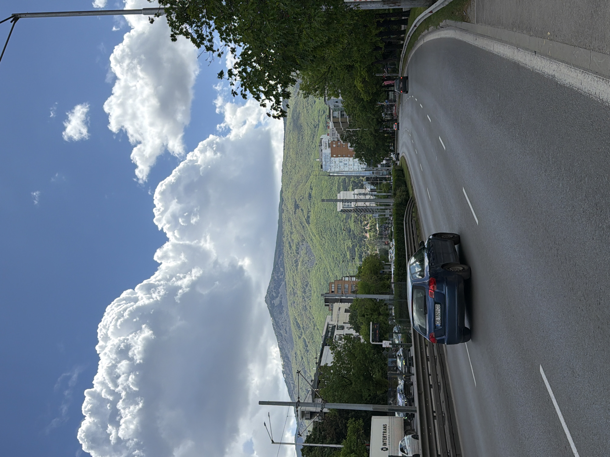 The image depicts a road with a blue car traveling on it. In the background, there is a mountain covered in greenery, with buildings situated at its base. The sky is filled with large, fluffy white clouds.
