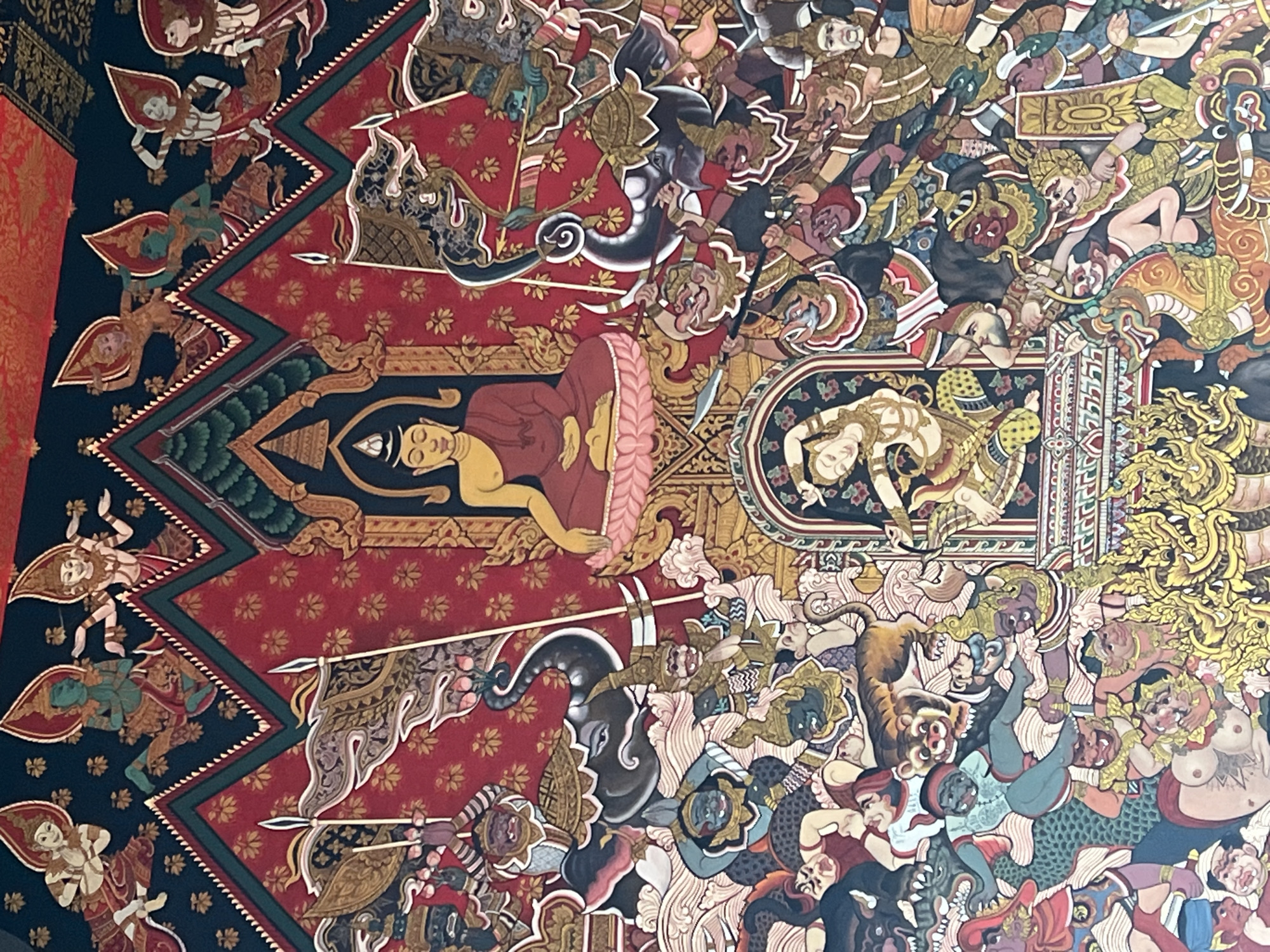 This image depicts a vibrantly colored mural with a central figure of Buddha seated in meditation. The mural is crowded with numerous figures, including deities, demons, and animals, engaged in a chaotic scene of conflict and movement. The style is reminiscent of traditional Thai art, with bold outlines and intricate details. The setting appears to be religious or cultural, possibly adorning a temple or public building. The location is identified as Baan Bo Phut, Thailand, suggesting it is a significant artistic representation within that locale.