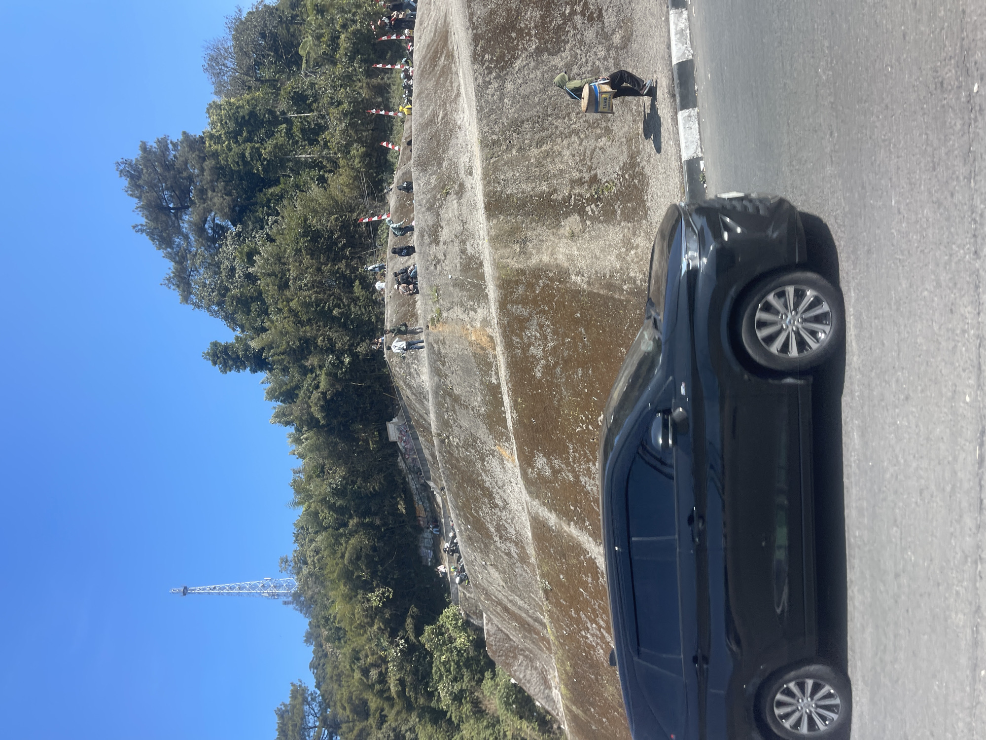 A black SUV drives along a paved road in front of a steep, terraced hillside. The hillside is covered in patches of green moss and brown dirt, with a concrete structure visible at the top. Several people are scattered across the terraces, some sitting and others standing. A person wearing a green hoodie and carrying a rectangular container on their back walks along the edge of the road. In the distance, a tall radio tower rises above a dense line of trees. The sky is clear blue. The scene appears to be the Puncak Pass road in Ciloto, Indonesia, a popular scenic route known for its hilly terrain and views.