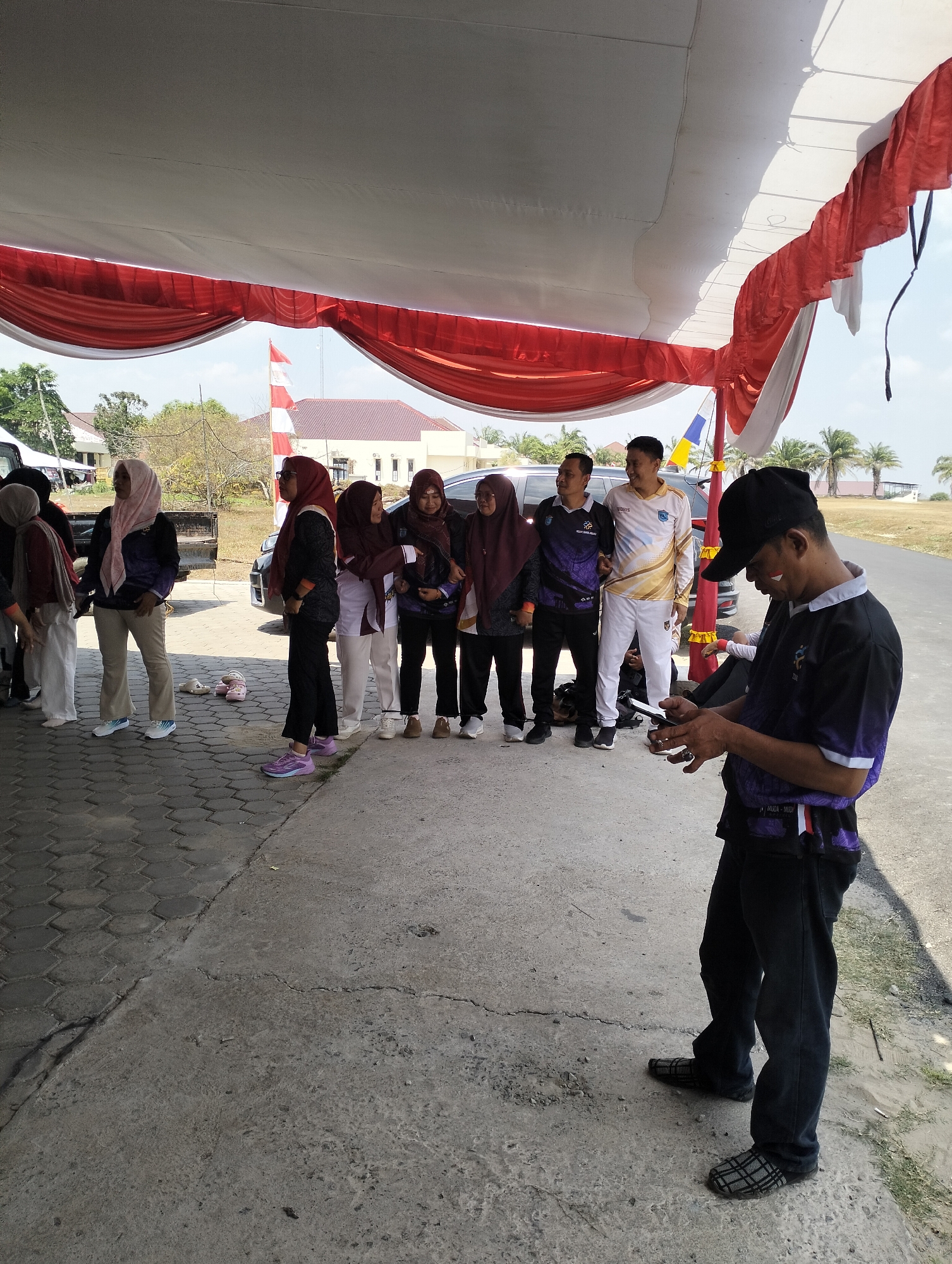 In Tanjung Sejaro, Indonesia, under a white and red tent, a group of people are gathered. A man in the foreground, wearing a black baseball cap and a purple and black shirt, is looking at his phone. Behind him, a line of individuals stand, some in hijabs and patterned shirts. Two men in athletic wear, one in white and gold, and another in purple, stand out. A car is visible in the background, along with palm trees and buildings. The ground is a mix of concrete and hexagonal pavers. The overall atmosphere appears to be an outdoor event or gathering.