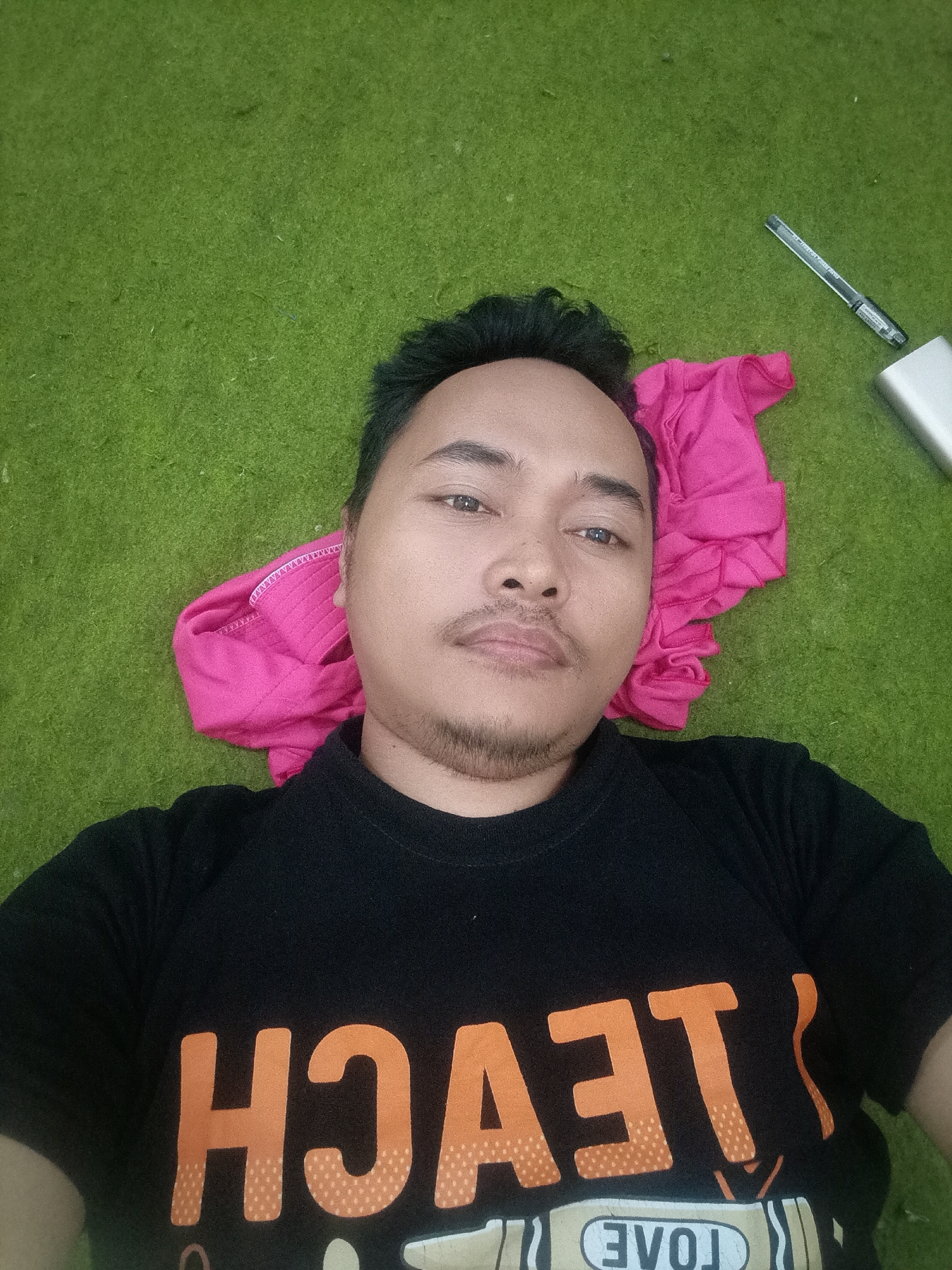 An overhead selfie shows a man lying on a green, carpeted surface, likely indoors. He is wearing a black t-shirt with orange lettering that partially reads "HOACH" and "LOVE". A pink garment is positioned beneath his head, and to his right, a silver power bank and a black pen are visible on the green surface. The man is looking directly at the camera with a neutral expression. The lighting suggests an indoor environment with artificial light. The overall scene is informal and casual. The image does not contain enough information to determine the specific location within Badas, Indonesia.