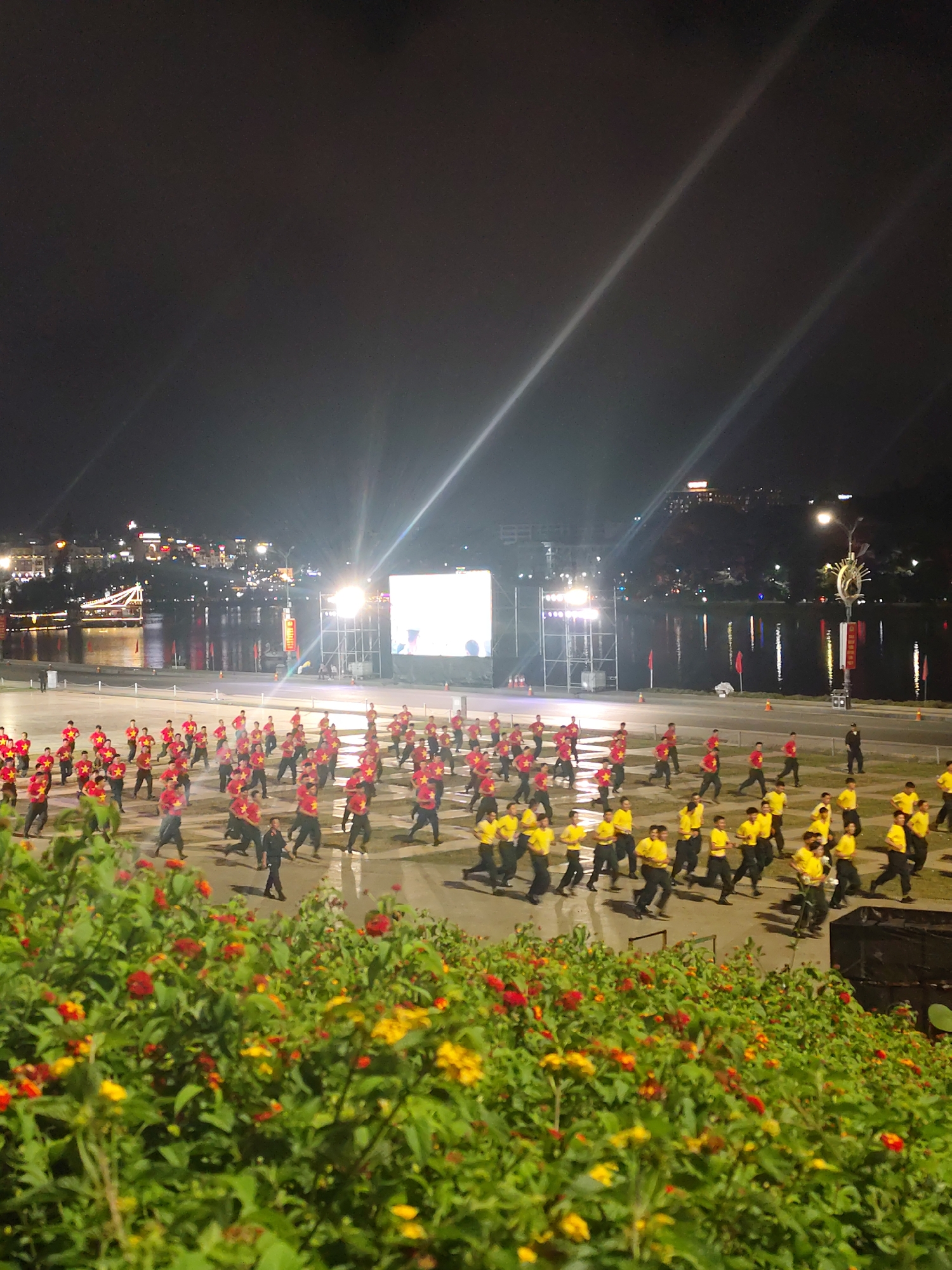 In Da Lat, Vietnam, a large group of people is participating in an outdoor event at night. The scene features a wide, paved area where hundreds of individuals are performing synchronized movements. The participants are mostly wearing red t-shirts with yellow stars, or yellow t-shirts. They are arranged in rows and columns, suggesting a choreographed performance or exercise routine.
In the background, a body of water is visible, reflecting the lights of buildings and structures along the waterfront. A bridge spans across the water in the distance. A large, illuminated screen is set up on a scaffolding structure, likely serving as a focal point for the event or broadcasting a performance. The surrounding area is lit by artificial lights, creating a dynamic nighttime atmosphere.
The time of day is clearly night, indicated by the darkness of the sky and the prominent artificial lighting. The weather appears clear, with no visible rain or fog. The presence of the water and the urban landscape suggests a waterside location within a city. The overall mood seems to be one of organized activity and public gathering, with a sense of energy conveyed by the synchronized movements of the participants. There is no visible text that can be deciphered in the image.
