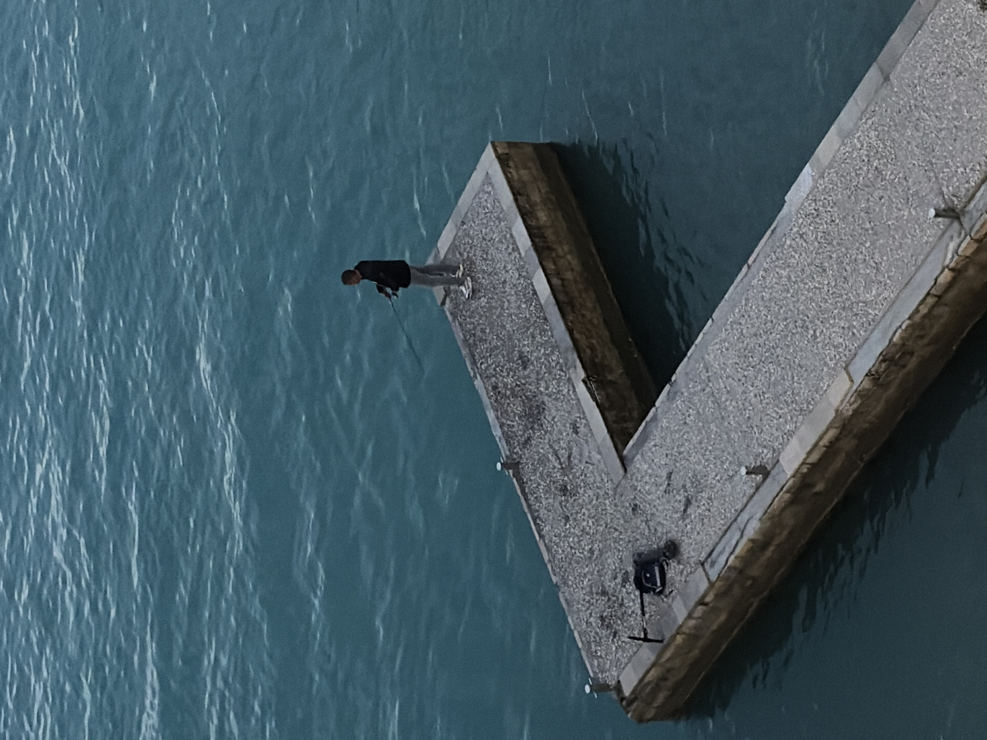 A man wearing a dark jacket and light-colored pants stands at the end of a V-shaped pier in the city of Budva, Montenegro. He is leaning over and appears to be looking at something in the water, possibly fishing with a fishing rod. A black bag or equipment case is present on the pier, along with metal posts. The surrounding water is a muted turquoise color with visible ripples.