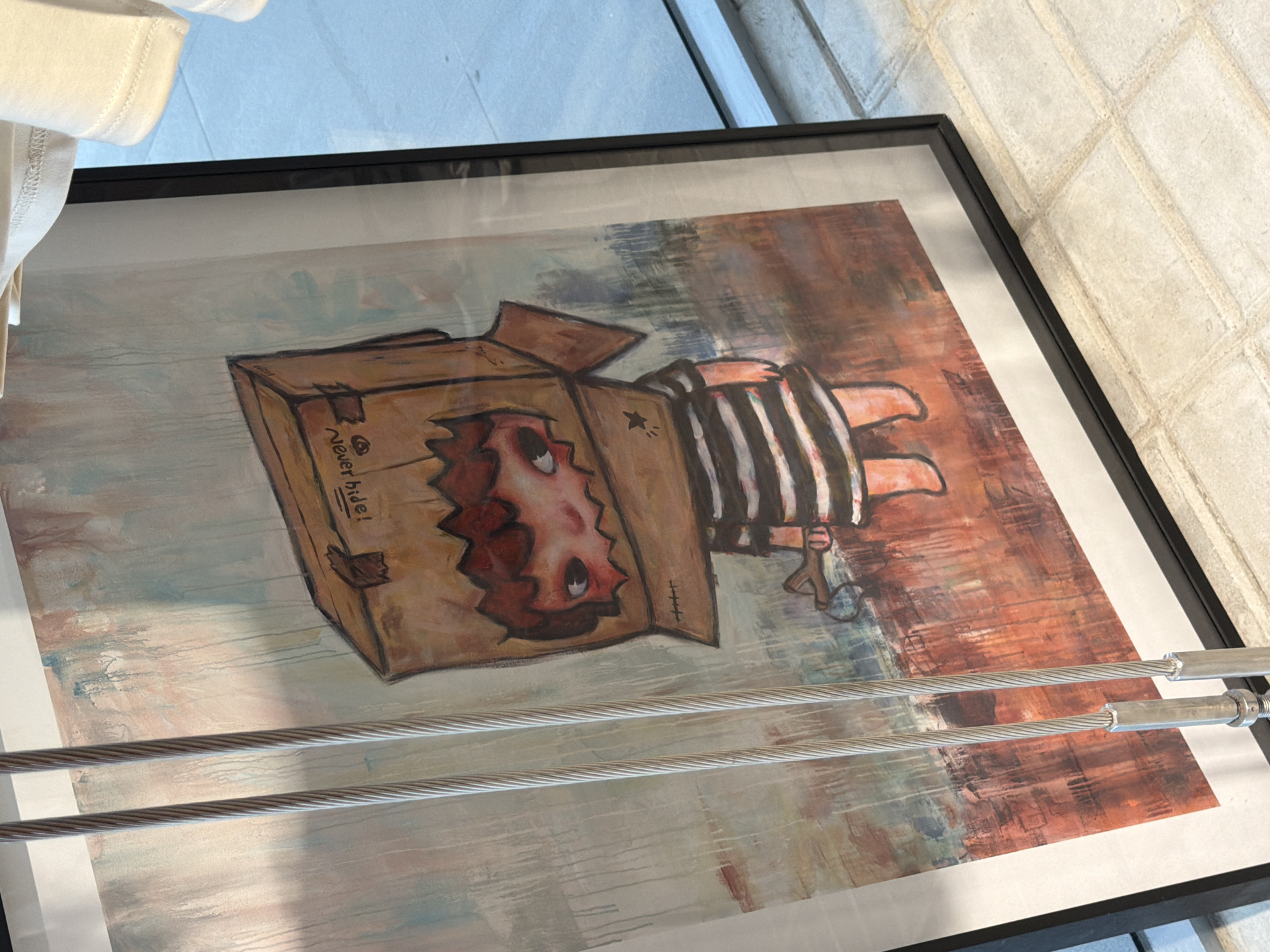 A framed painting is displayed in a well-lit indoor space with a tiled floor. The painting depicts a cartoonish child wearing a cardboard box over its head. The child has large, expressive eyes peeking out from cutouts in the box, and is wearing a striped shirt. The background of the painting features abstract brushstrokes in various colors. Two metal cables hang in front of the painting, partially obscuring it. A white garment, possibly a t-shirt, is visible at the top right of the image. The overall setting appears to be a retail environment, possibly a boutique or gallery, given the display of merchandise. There are no people actively interacting with the painting. The presence of a white garment and the general presentation suggest a shopping or exhibition context in a city like Bangkok, Thailand, where such displays are common in trendy areas.