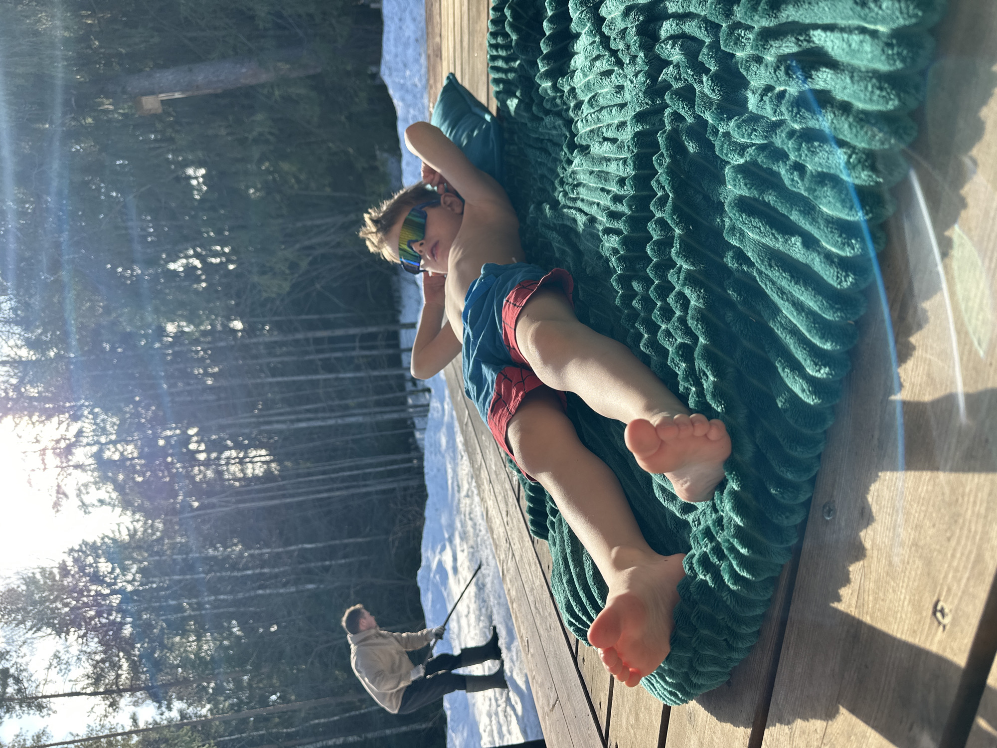 A daytime outdoor scene in Прикамский, Russia, depicts two males on a wooden deck adjacent to a snow-covered area and a dense evergreen forest. In the foreground and midground, a young male child is lying shirtless on his back on a teal-colored, textured blanket, resting his head on a small matching pillow. He wears blue swim trunks with red accents and multi-colored reflective sunglasses, with his hands positioned behind his head and bare feet visible. In the background, to the left, an adult male is standing on a patch of snow-covered ground, facing away from the camera and slightly right. He is dressed in a light-colored jacket, dark trousers, and dark rubber boots, holding a long-handled tool, possibly a shovel, engaged in an activity on the snow. The background is dominated by a dense forest of tall, slender trees. Bright sunlight illuminates the scene from the upper right, casting shadows and causing lens flare. A small wooden birdhouse or similar structure is visible on a tree trunk in the upper right background.