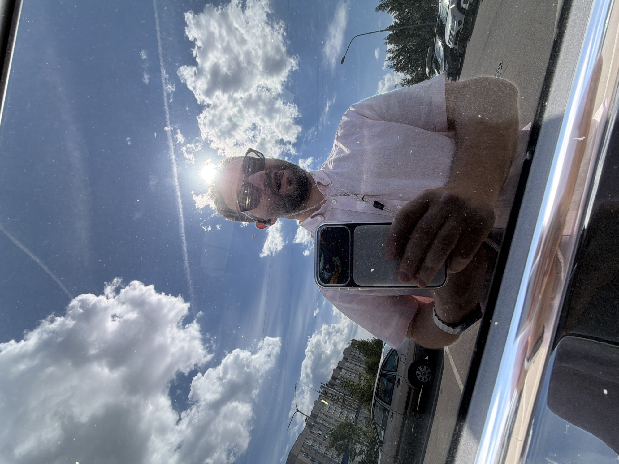 The image is a selfie captured in a reflective surface, likely a car window, showing a Caucasian male in his 30s-40s with a beard, sunglasses, and wired in-ear monitors. He wears a light pink short-sleeved collared shirt and a silver watch on his left wrist. His left hand holds a dark smartphone, possibly an iPhone, with a visible camera module and case. The man's mouth is slightly open.
The background, visible through the reflection, depicts a sunny urban environment. The sky is a clear blue with large, scattered white cumulus clouds and two distinct airplane contrails. A bright sun, positioned behind the man's head, creates significant glare and lens flare. On the left, a multi-story residential apartment building with a light-colored facade, characteristic of post-Soviet architecture, is visible. Parked cars include an older silver sedan on the left and several modern vehicles, including an SUV with a roof rack, in a parking lot on the right. Green trees are interspersed between buildings and vehicles, and a dark street lamp pole stands on the right. The reflective surface itself shows numerous small bright speckles, indicative of dust or minute imperfections. This scene is consistent with a typical residential area within the city of Saint Petersburg, Russia.
