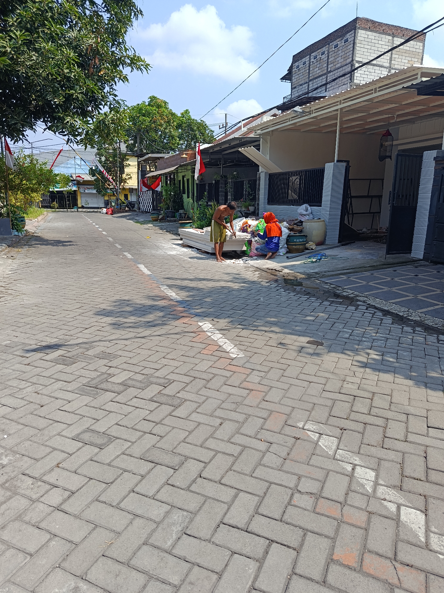 A paved street in Keboananom, Indonesia, lined with residential buildings and trees, is captured under a bright sky with scattered clouds. On the right side of the street, two individuals are engaged in an activity near a pile of white materials, possibly construction supplies. One person, wearing a bright orange head covering and blue clothing, is seated and interacting with the materials. Another person, wearing a yellow loincloth, stands and appears to be assisting or observing. In the background, several houses with varying architectural styles are visible, some adorned with Indonesian flags. The street itself is paved with interlocking gray bricks, with a white dashed line marking the center. Shadows from the surrounding structures and foliage are cast across the pavement. A birdcage hangs from an overhang on the right side of the image.