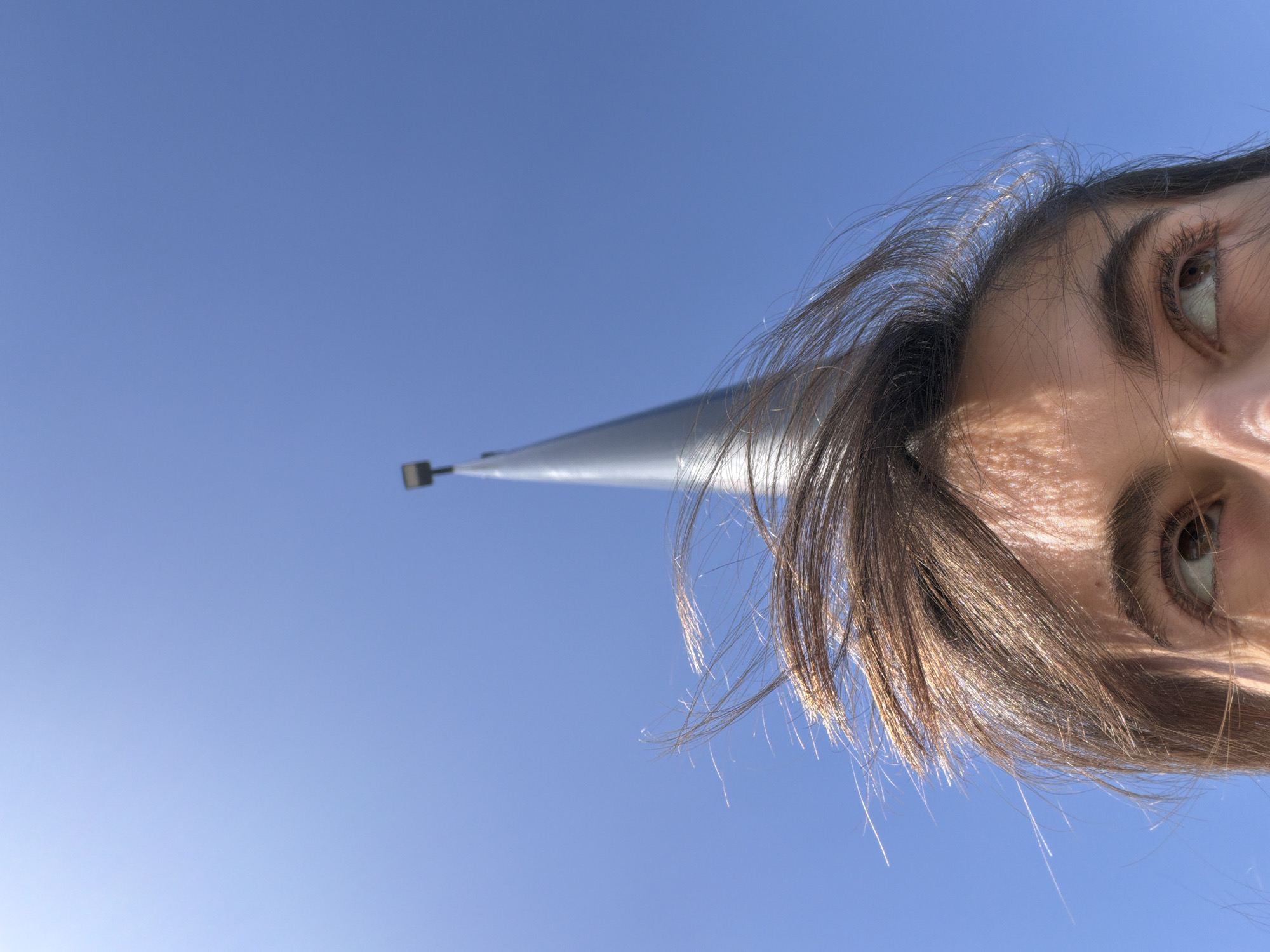 The image captures a low-angle perspective looking upwards towards a clear blue sky. In the foreground, on the right side of the frame, is a close-up, partial view of a young person's face. Visible are their right eye, eyebrow, forehead, and a mass of dark brown hair, illuminated by bright sunlight. The person's eye is open and looking towards the top of the frame. The sunlight creates highlights on the hair and subtle shadows and texture on the skin.
Extending diagonally from the left side of the frame, behind the person's head, is a slender, tapering metallic pole, likely silver or grey. A small, dark, rectangular object is visible at the very top of the pole. The sky is uniformly blue, indicating clear, sunny weather during daytime. The image is presented rotated 90 degrees clockwise from an upright orientation. There is no visible text in the image. The scene is outdoors, set in Mascouche, Canada.