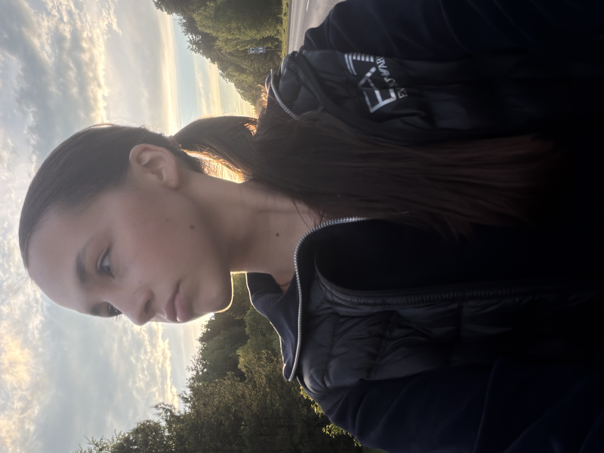 A young woman with dark hair in a ponytail poses in an outdoor setting. She wears a dark-colored vest over a navy-colored sweater. The sky is visible in the background and is partially obscured by trees. The setting appears to be near a road with trees lining the background. The location is in Chaikovsky, Russia.