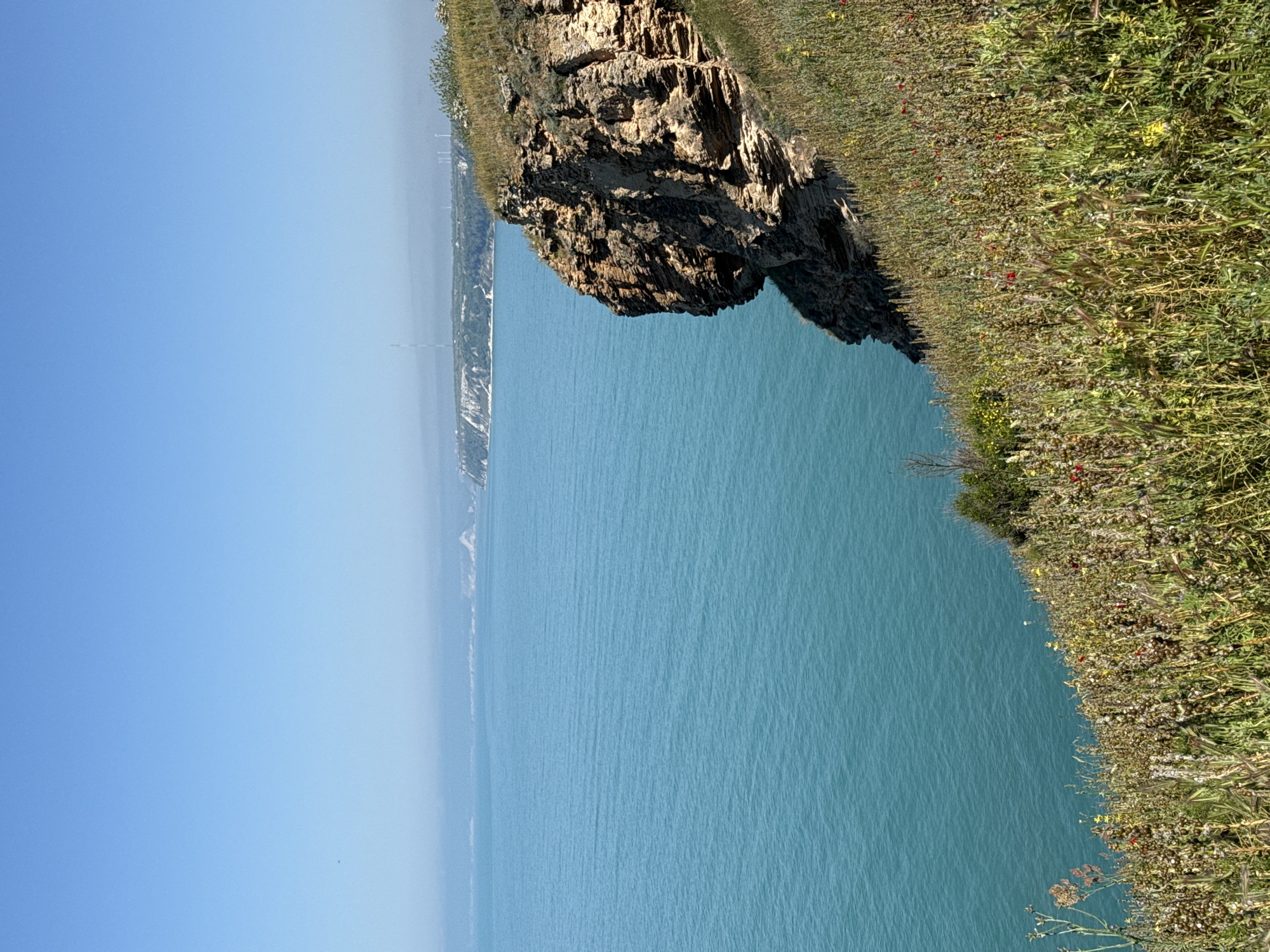 The image depicts a coastal landscape in Balgarevo, Bulgaria. A rocky cliff covered with greenery overlooks the blue sea. The sky is clear and bright. In the distance, a stretch of land is visible along the horizon, appearing hazy. There are no people or visible interactions in the frame. The focus is on the natural scenery.