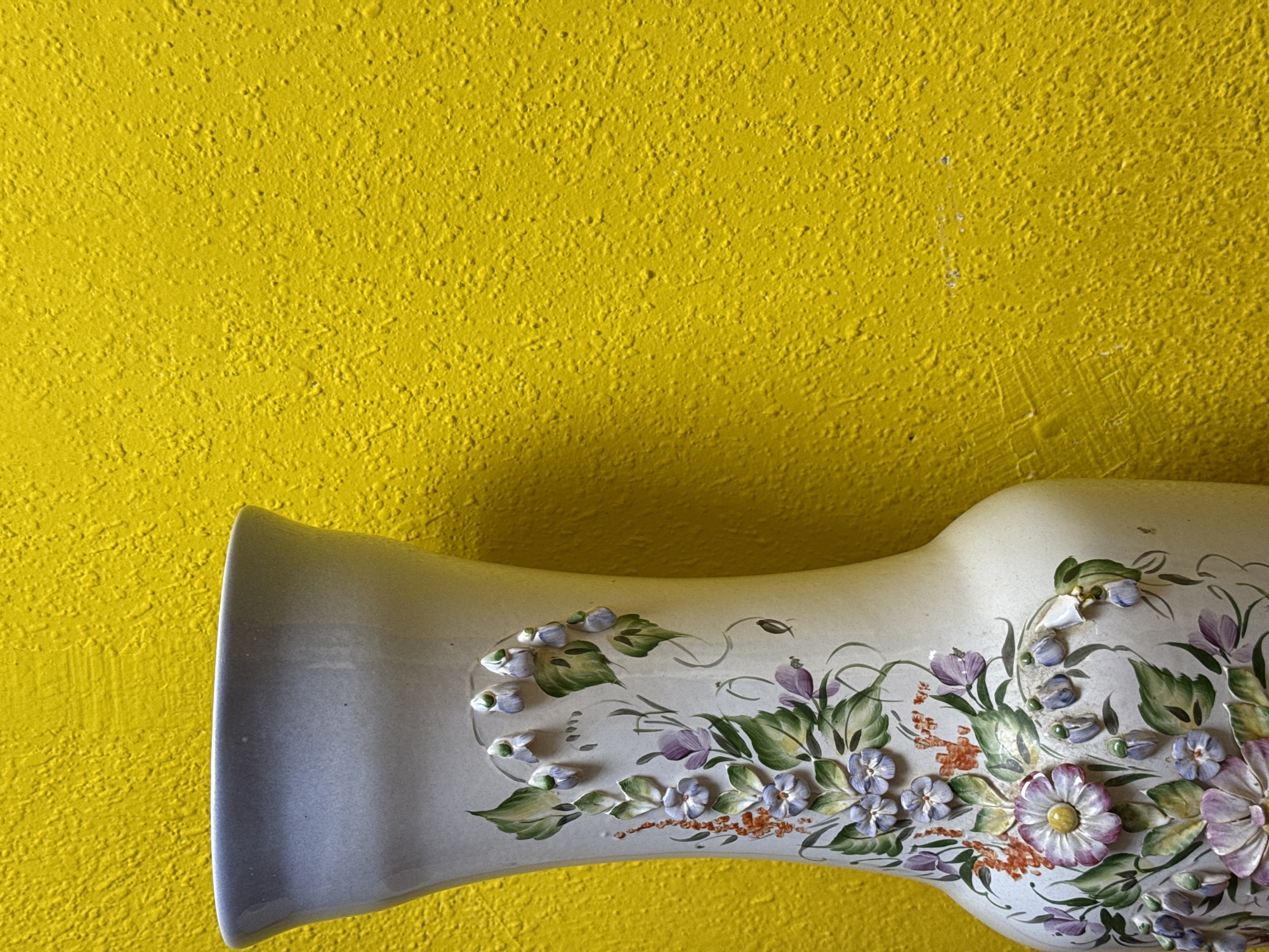 A cream-colored vase decorated with raised floral designs rests against a heavily textured yellow wall. The vase is horizontally oriented, with the flared opening on the left. The floral patterns include green leaves and purple and white blossoms.