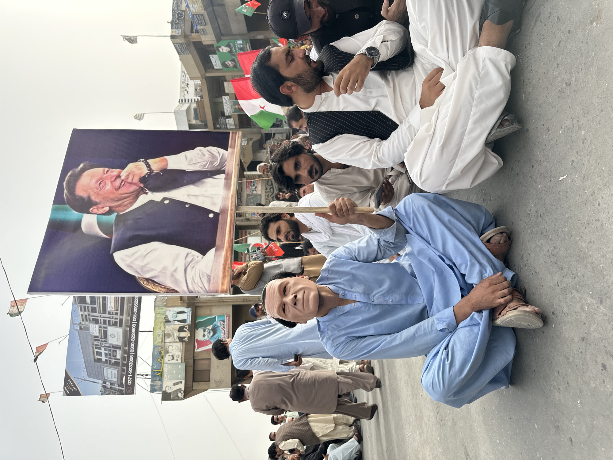 A crowd of men is gathered outdoors on a paved surface, likely in Quetta City Tehsil, Pakistan. In the foreground, two men are seated on the ground. The man on the left wears a light blue shalwar kameez and a mask that resembles the face of a Caucasian man. He is holding a wooden pole. The man on the right wears a white shalwar kameez with a black pinstriped vest. He is looking down at his hands, one of which has a watch on it. Behind them, other men are standing and seated. A large banner featuring a smiling man in a dark vest over a white shirt is prominently displayed. Several flags, including red, white, and green ones, are visible throughout the scene. Buildings with various signage and posters are in the background. The sky is overcast.