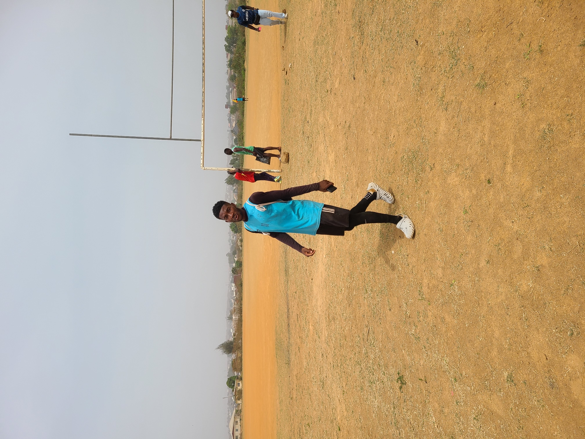 The image depicts an individual standing on a dry, grassy field in Barikin Jirga Summa, Nigeria. The person is wearing a light blue shirt, black shorts, and white sneakers. In the background, there are other individuals and a rudimentary goalpost. The scene suggests a sports field or recreational area within the city. The sky is overcast.