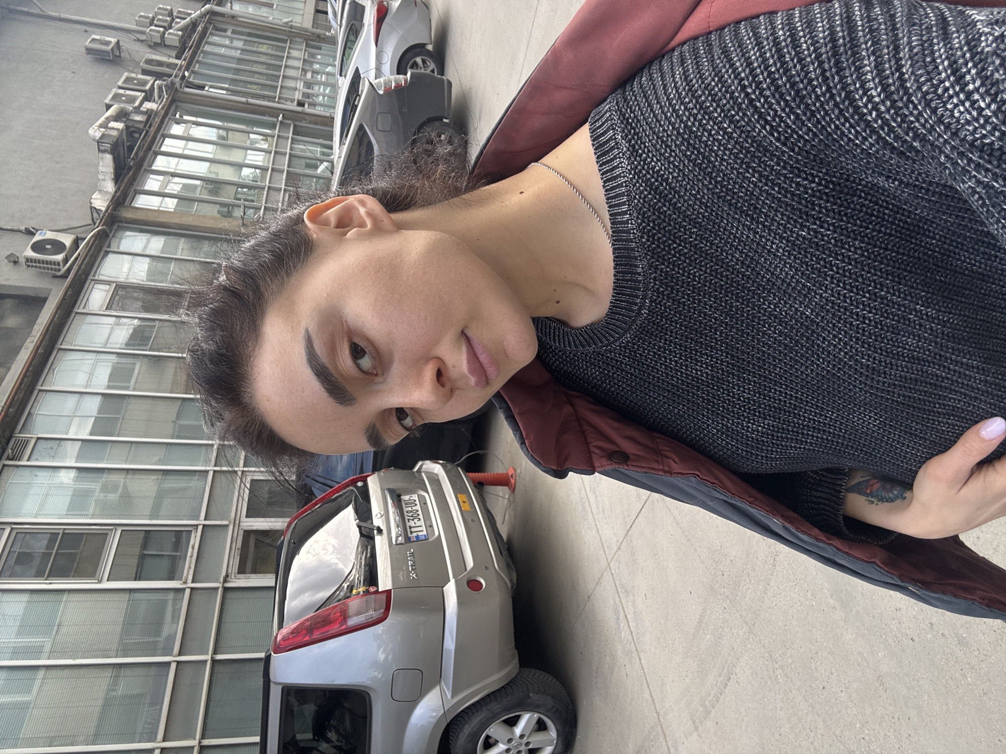 A close-up selfie shows a person from the chest up, looking directly at the camera with a neutral expression. The person has dark hair pulled back and is wearing a dark, possibly charcoal or navy, knitted sweater layered under a dark red or maroon jacket. A thin silver chain is visible around their neck. On their left inner forearm, a colorful tattoo, possibly a butterfly, is visible. Their left hand, with light-colored nail polish, rests near their hip.
The background reveals an urban setting, with the person positioned near parked cars and a multi-story building. Directly behind the person is a light gray Nissan X-Trail SUV, displaying a Georgian license plate "TT-368-JU" and the "X-TRAIL" badge. A red traffic cone is partially visible behind the SUV. Another gray car and a white car are parked further back. The building in the background features numerous windows with thin frames and several exterior air conditioning units mounted on its facade, suggesting an older commercial or industrial structure. The scene is brightly lit, indicating it is daytime, possibly overcast, given the soft light and reflections in the windows. The overall setting points to a street or parking area in Tbilisi, Georgia, as indicated by the license plate and provided context.