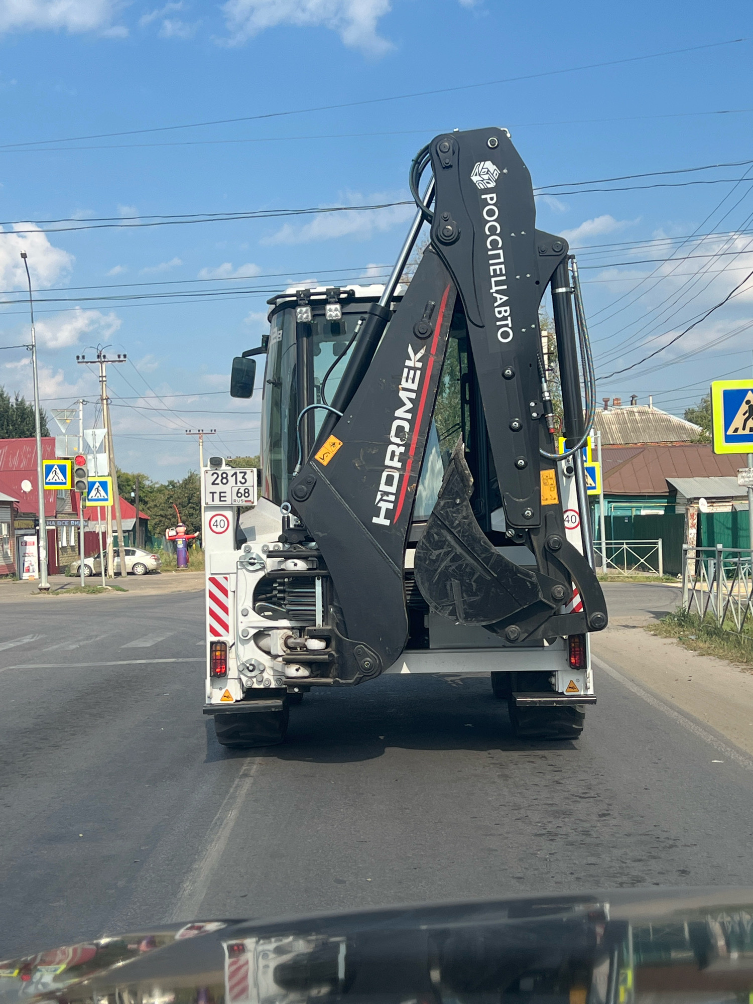 The image shows the rear view of a white and black backhoe loader, with the brand name "HIDROMEK" prominently displayed in large white letters on the black arm. The loader is parked on an asphalt road, likely at an intersection, given the presence of traffic lights and pedestrian crossing signs in the background. The sky is blue with scattered clouds.
To the left of the loader, a traffic light with red and yellow signals is visible, along with two yellow pedestrian crossing signs. Further in the background, there are several residential buildings with red and brown roofs, surrounded by trees. A white car is parked near one of the buildings. On the loader itself, a license plate reading "2813 TE 68 RUS" is affixed, along with a circular sign indicating a speed limit of 40.
The inscription "РОСПЕЦПРОМ" is visible on the loader's arm. The overall scene suggests a daytime setting in a moderately urban or suburban area of Моршанск, Russia, with the backhoe loader appearing to be stationary.